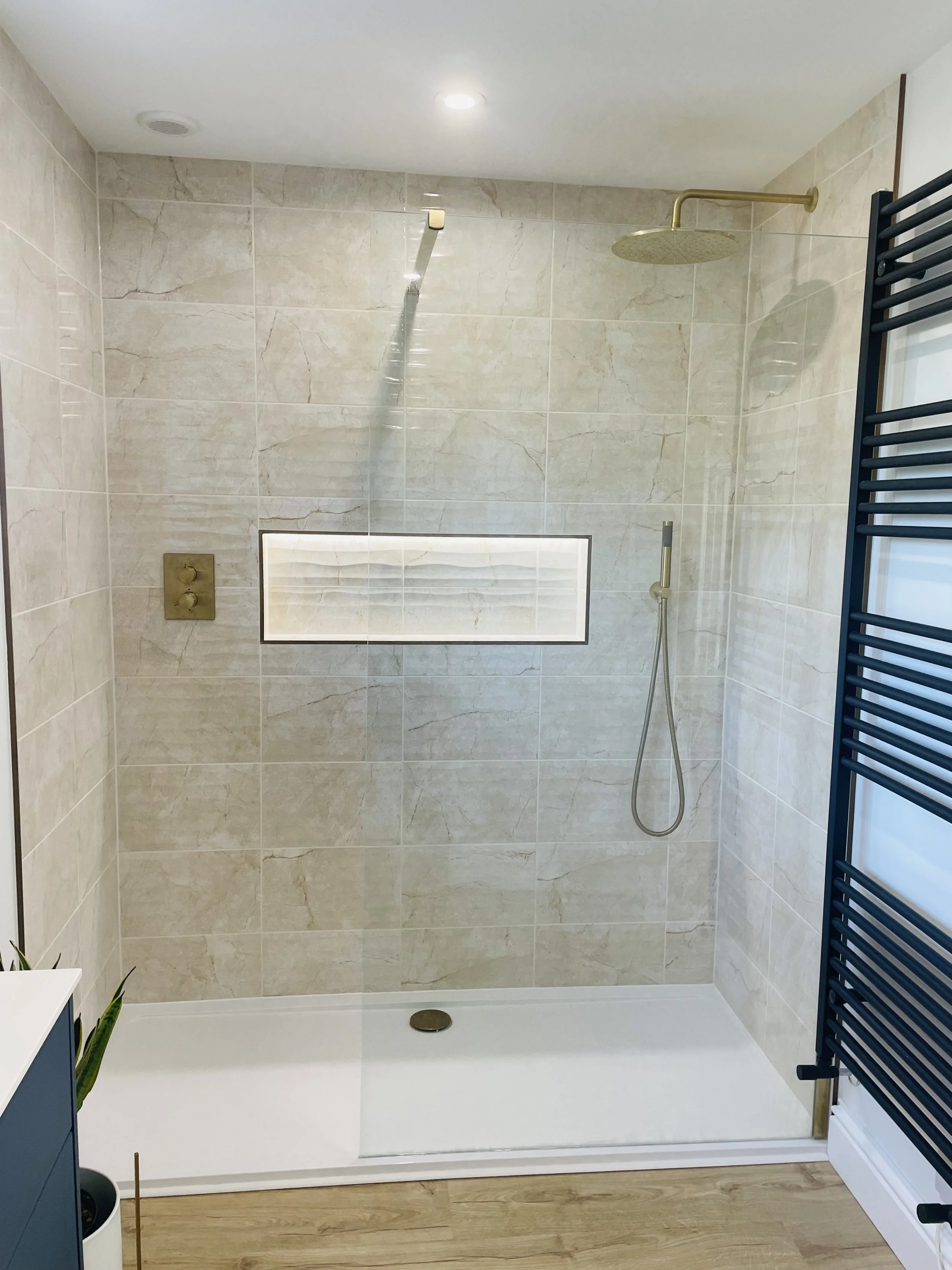 Modern walk-in shower with beige marble tiles, a gold rain showerhead, a handheld shower wand, a built-in niche for toiletries, and a black heated towel rack.