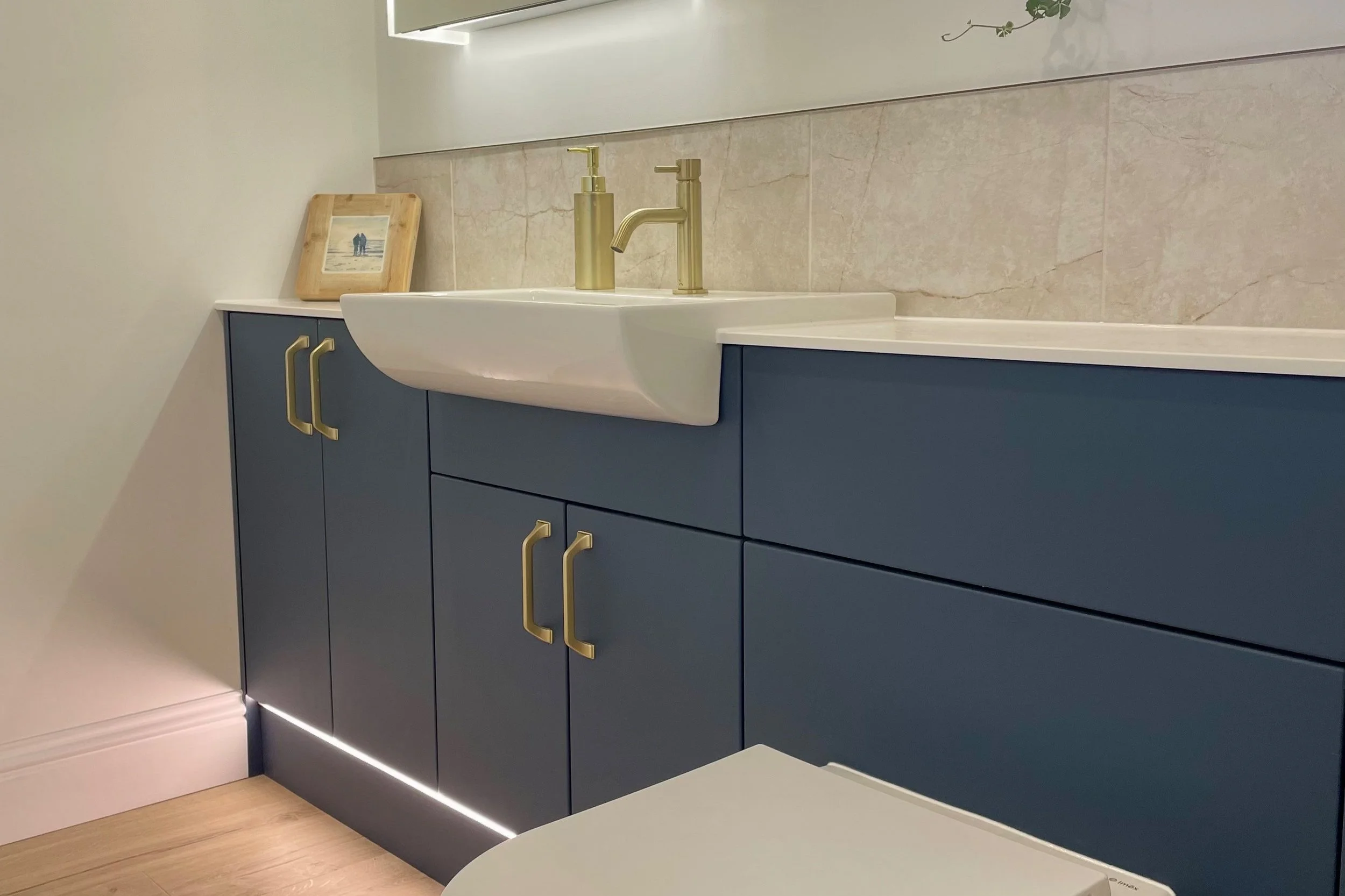 Close-up view of a modern bathroom vanity with blue cabinets, a white countertop, a small white sink, and gold faucet fixtures. A framed picture with a beach scene rests on the countertop, and there's a marble tile backsplash. Part of a white toilet is visible in the foreground.