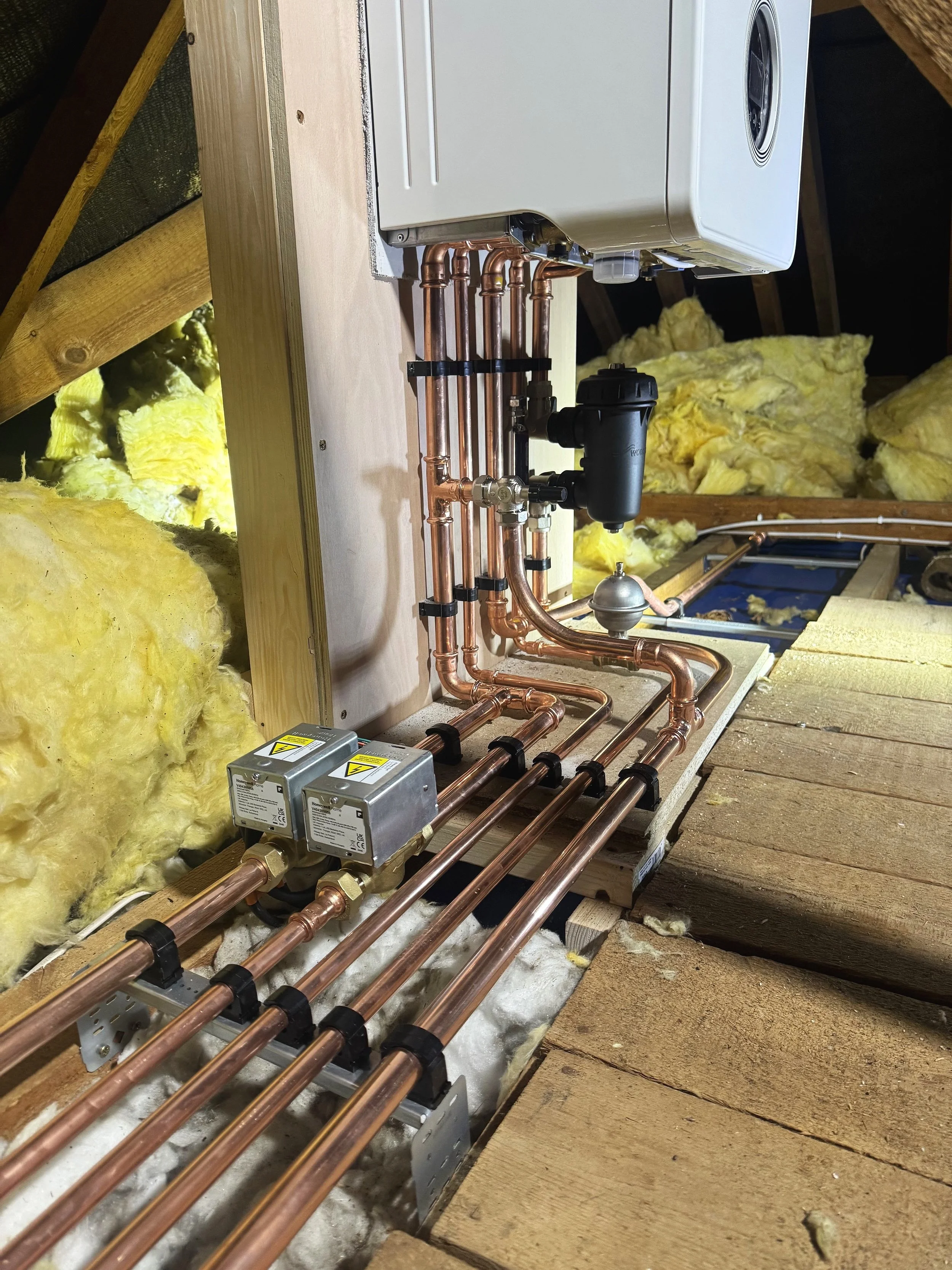 Piping system of copper pipes connected to a water heater, installed in an attic with insulation and wooden beams.