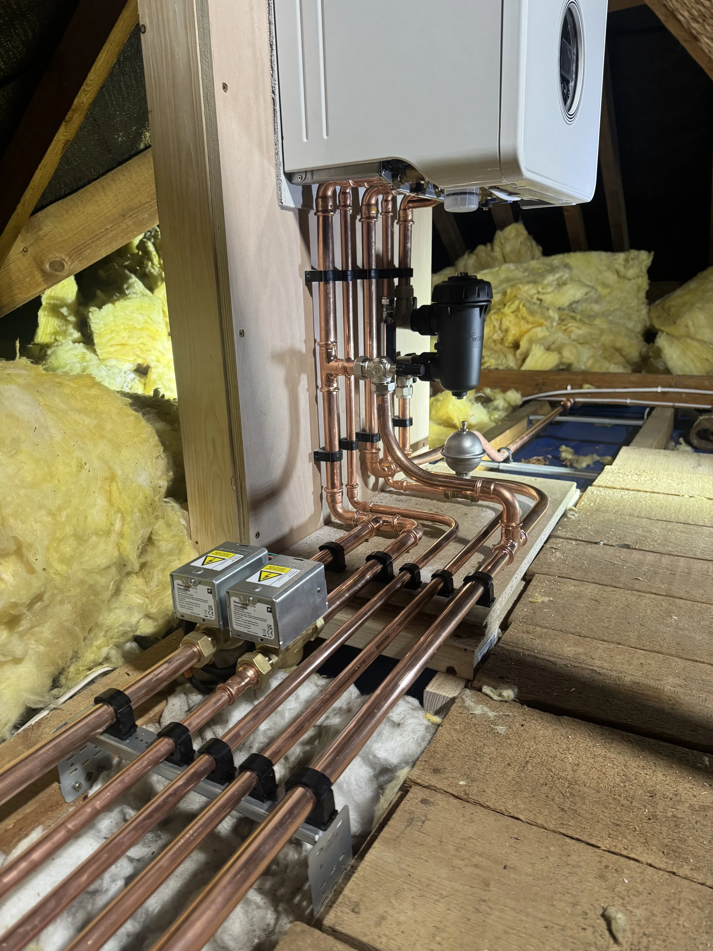 Copper piping and a white washing machine installed in an attic with insulation and wooden floorboards.