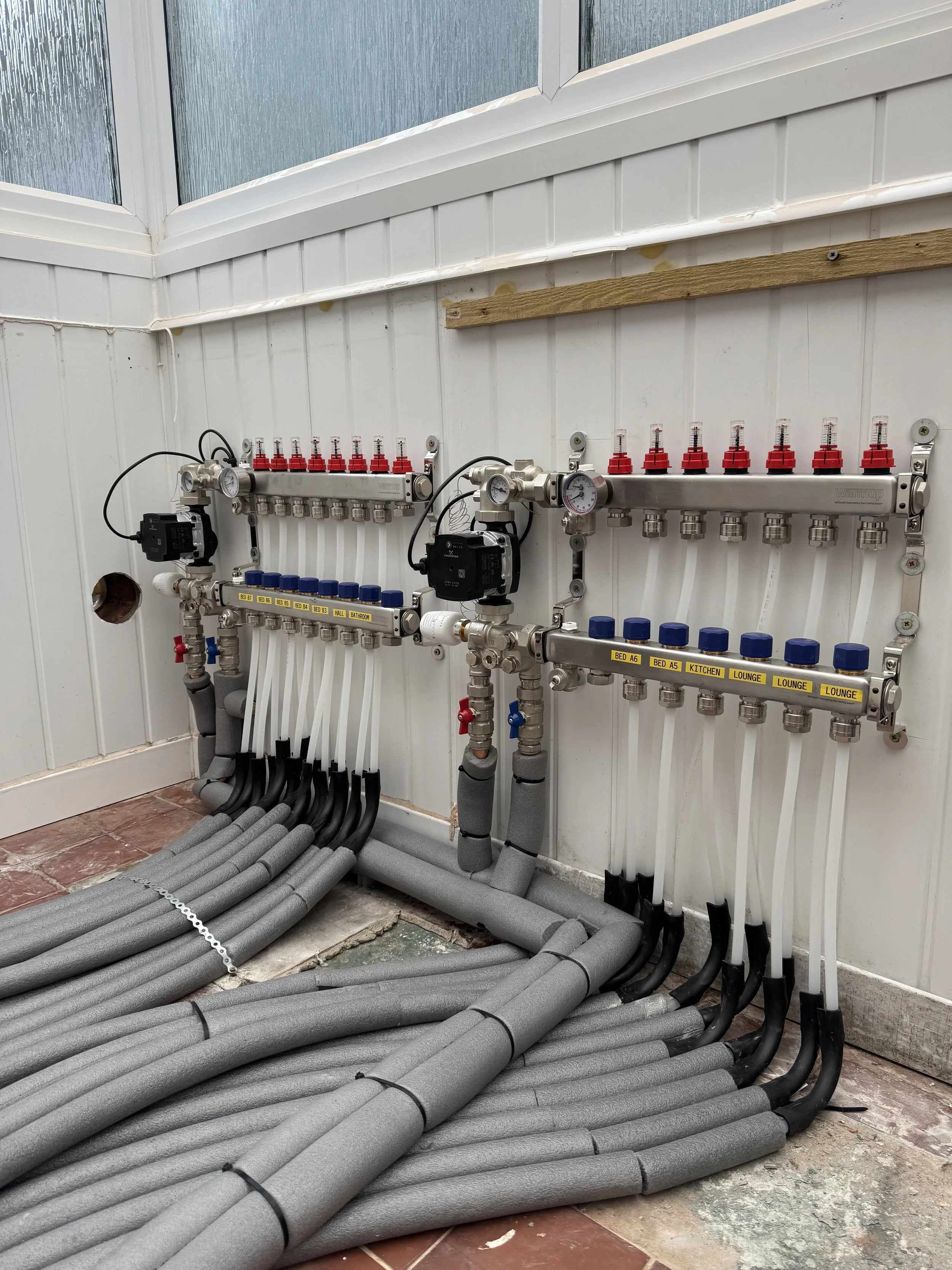 A plumbing manifold system with multiple pipes, valves, and gauges installed on a wall, with foam insulation on the pipes on the floor.