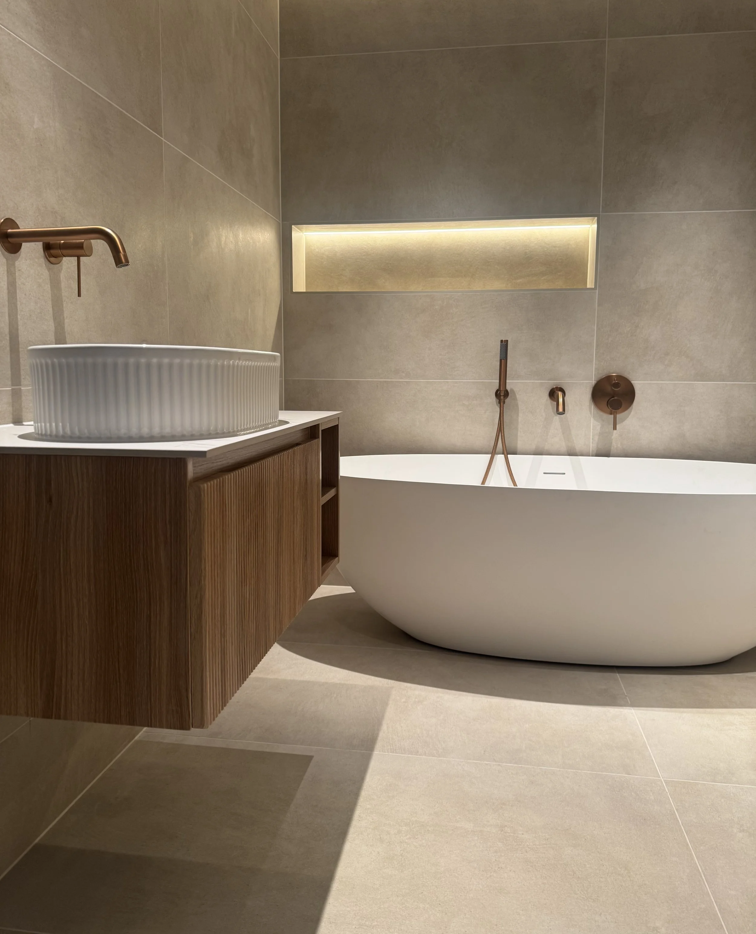 Modern bathroom with beige tile walls and flooring, a white freestanding bathtub with copper fixtures, a wooden vanity with a round ribbed white vessel sink, and a rectangular recessed light above the bathtub.