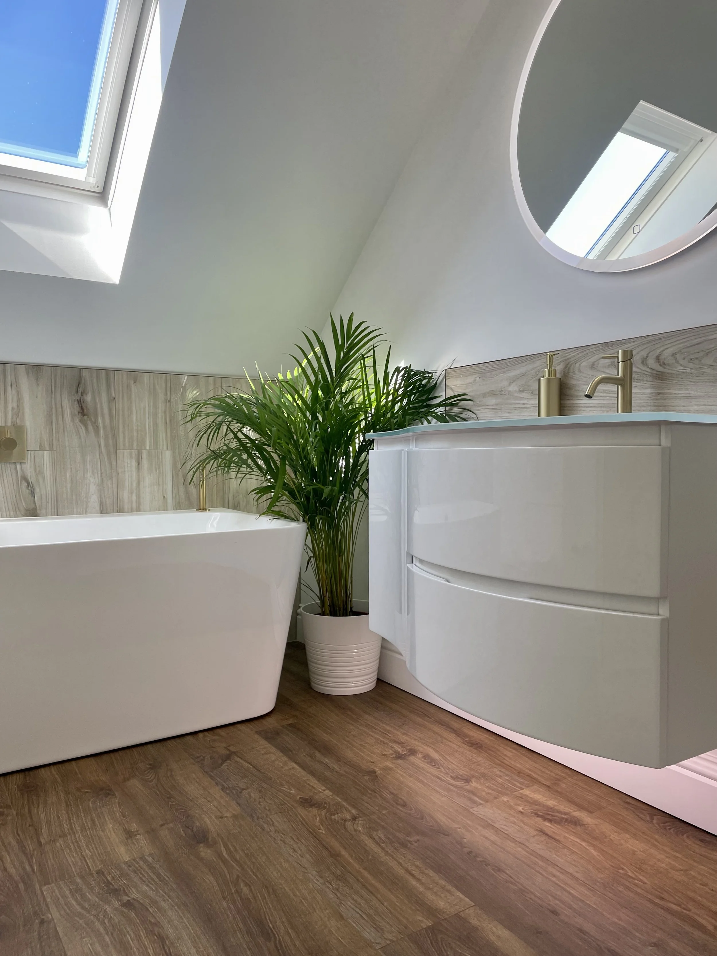 A modern bathroom with a white freestanding bathtub, a white vanity with gold fixtures, a large green potted plant, a wooden floor, and a skylight window on the sloped ceiling.