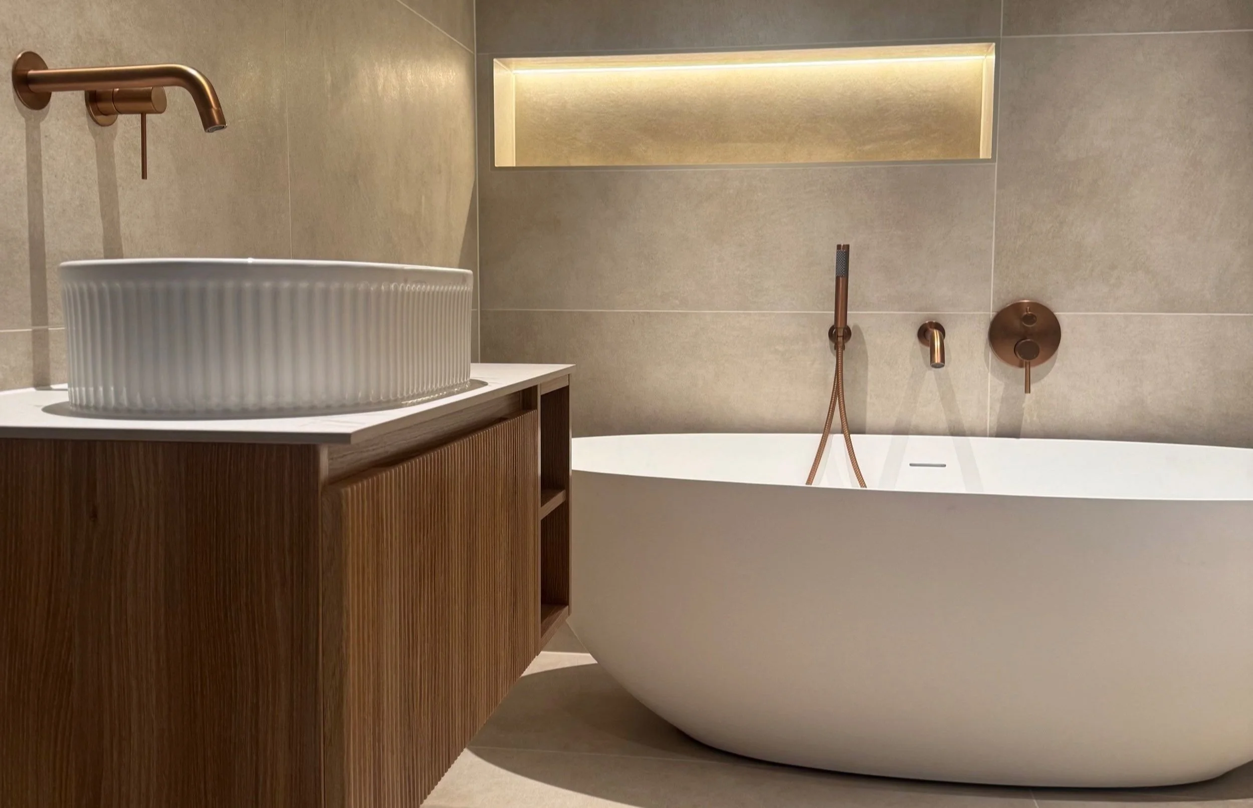 Modern bathroom with a freestanding white bathtub, a white ribbed vessel sink on a wooden vanity, and brushed bronze fixtures. There is a rectangular niche with lighting in the beige tiled wall.