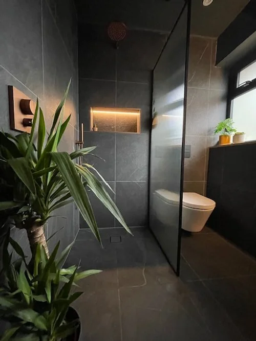 Modern bathroom with dark gray tiles, a floating toilet, a glass shower partition, and green plants near the window.