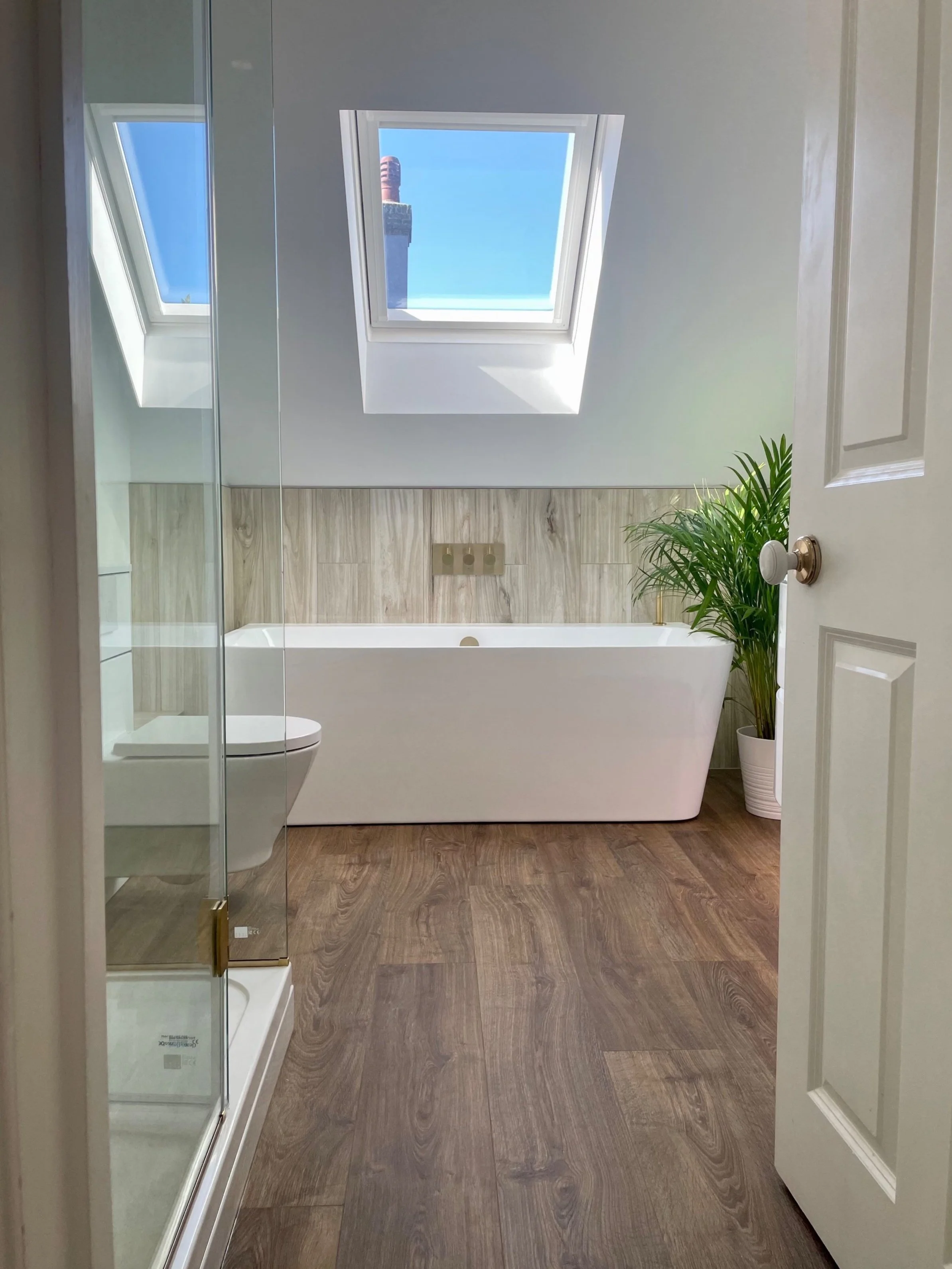 Modern bathroom featuring a bathtub under a skylight, wooden flooring, a plant in a white pot, and a partial view of a shower enclosure and toilet.
