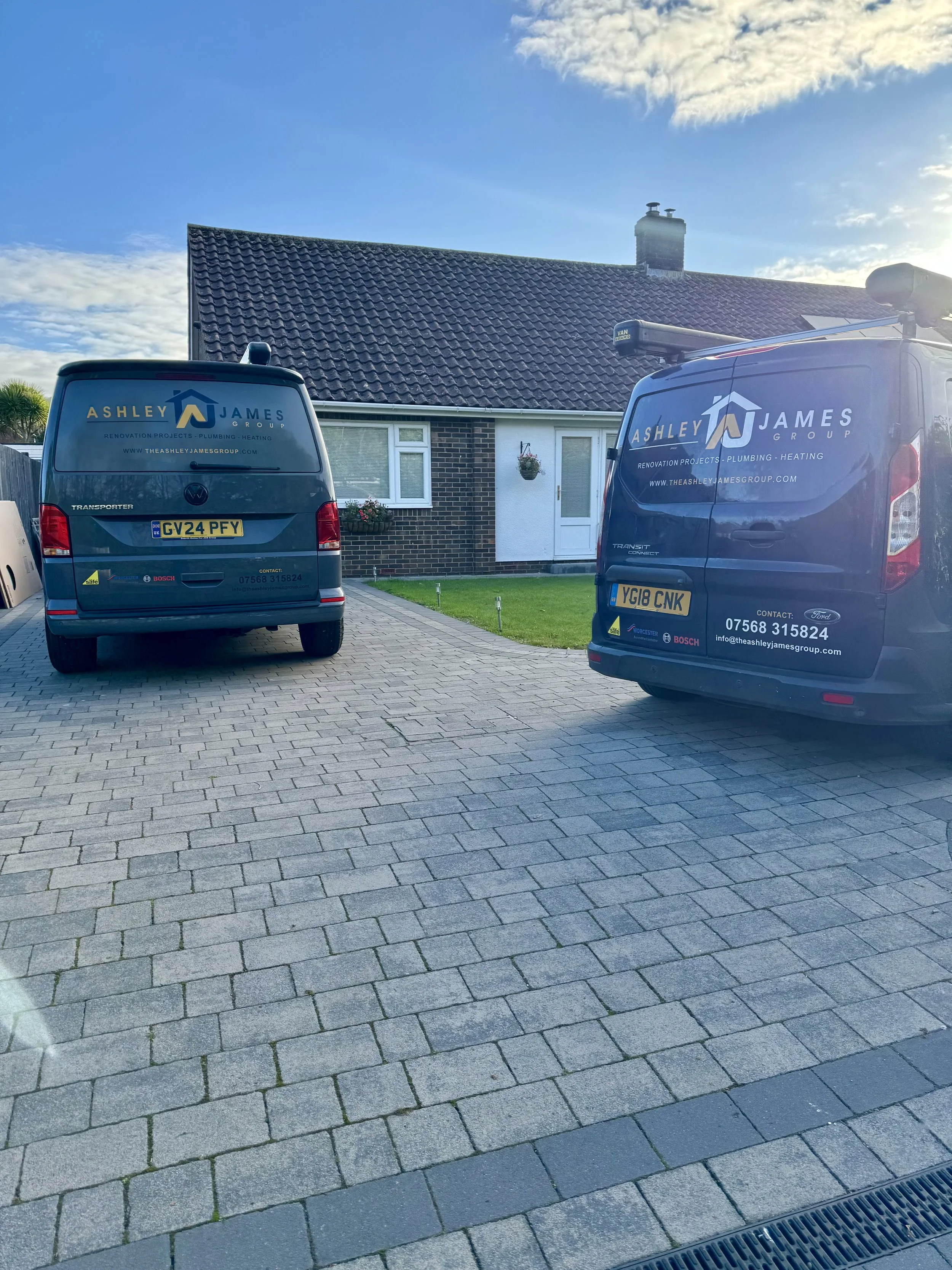 Two Ashley James Group service vans parked in front of a house with a grey tiled roof, brick walls, and a small front lawn, under a blue sky with some clouds.