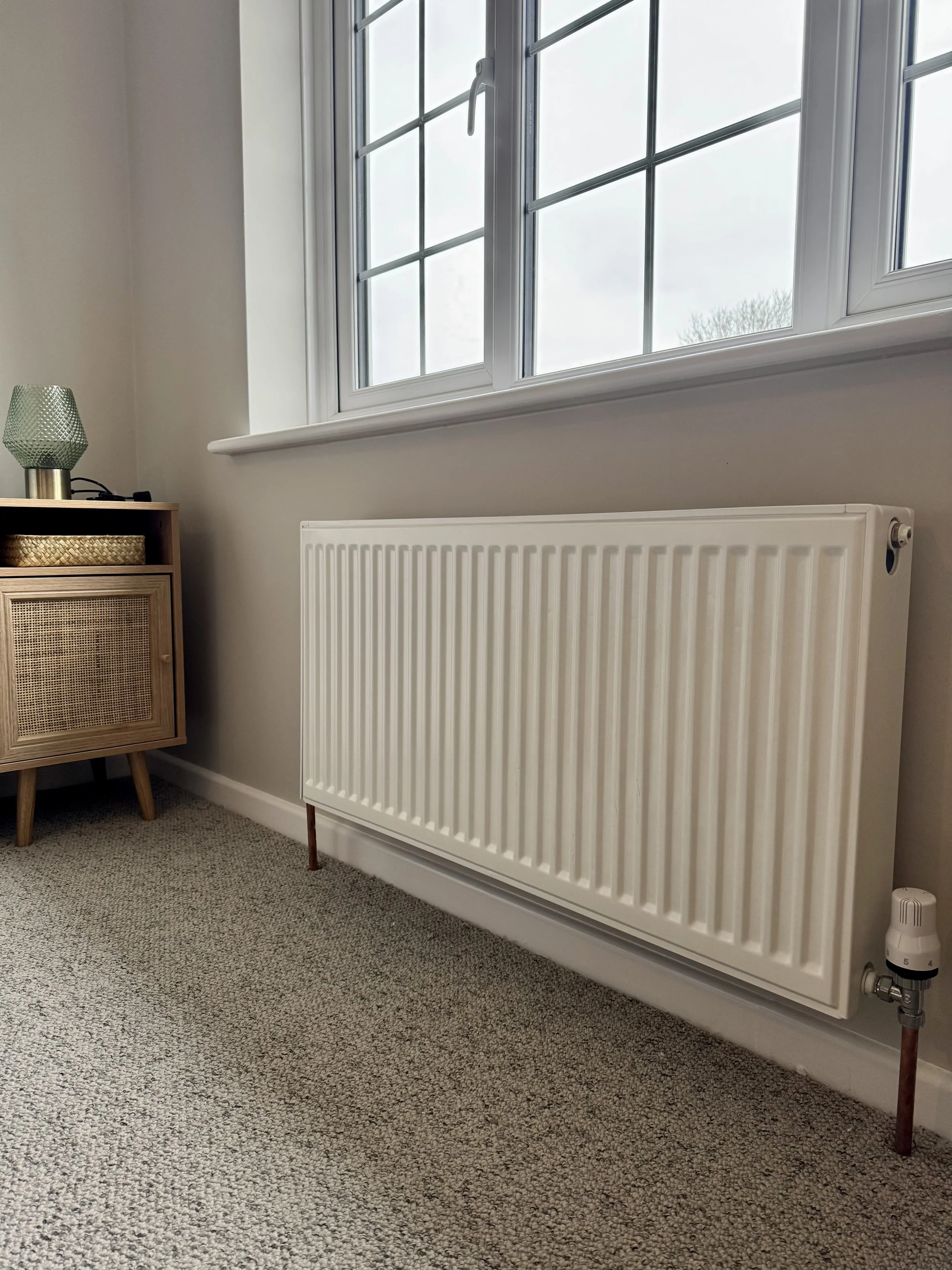 A white radiator mounted on a beige wall beneath a window in a room with beige carpet and a wooden side table with a green lamp.