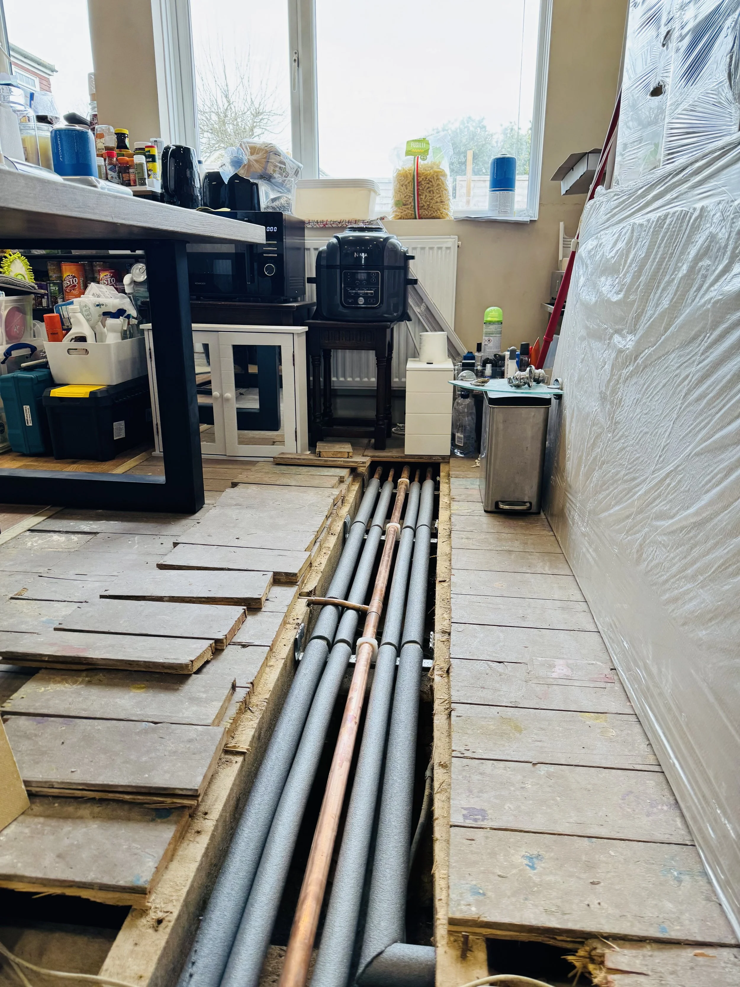 Room with exposed floor showing electrical and plumbing pipes, partially tiled floor, cluttered with tools, appliances, shelves, and a large window.