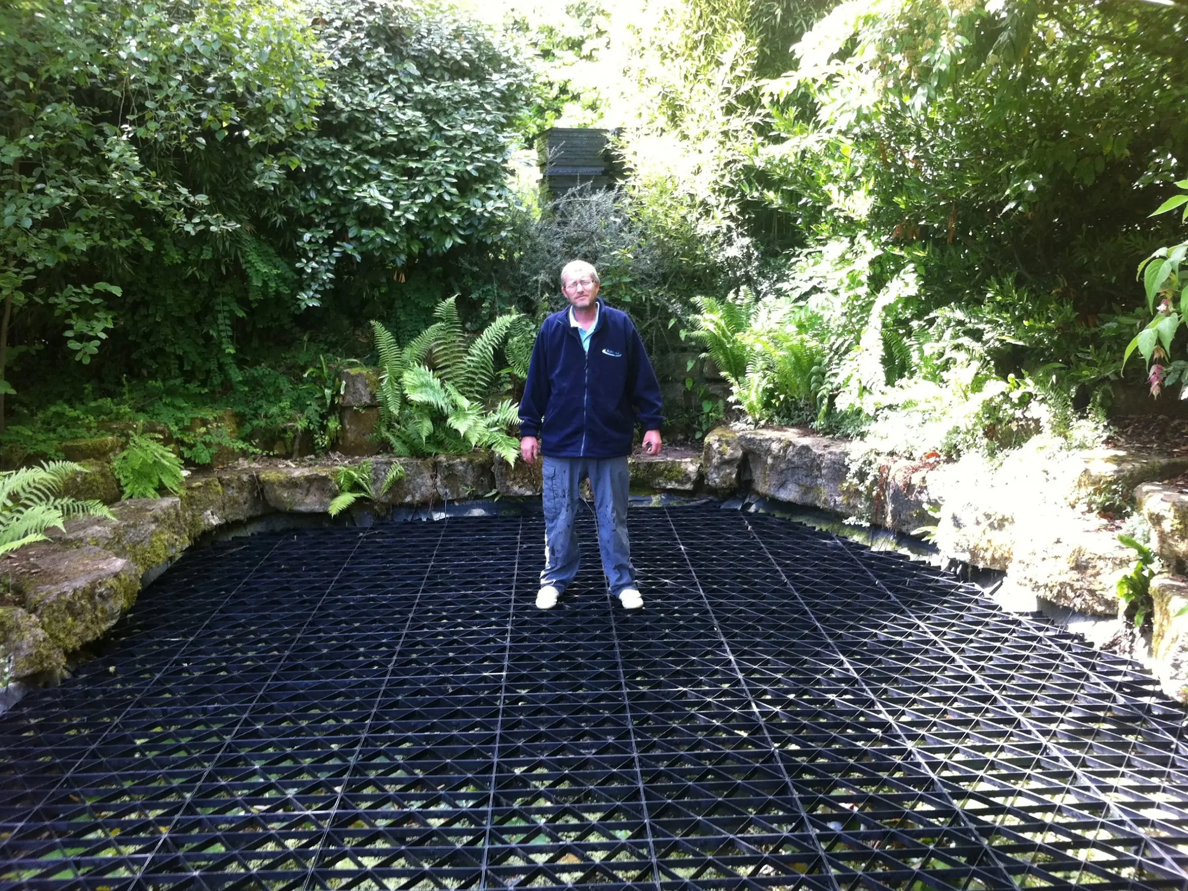 A man standing on a black, grid-like surface in a lush garden with green foliage and ferns.