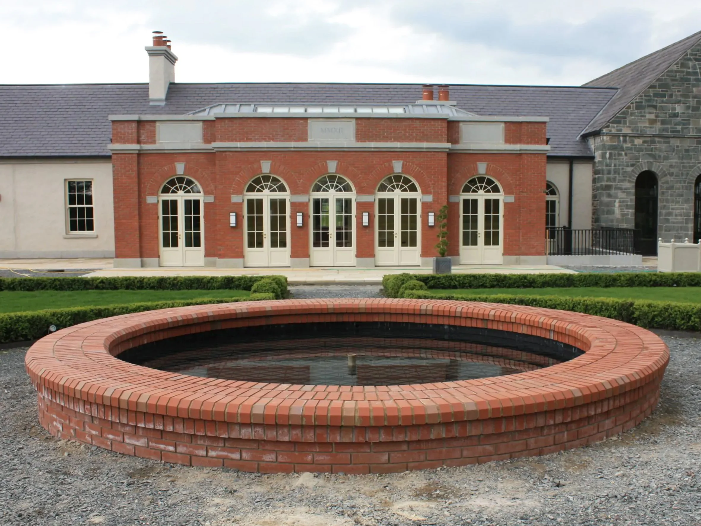 A circular brick fountain in the foreground with a historic brick and stone building with large arched windows and a pitched roof in the background.