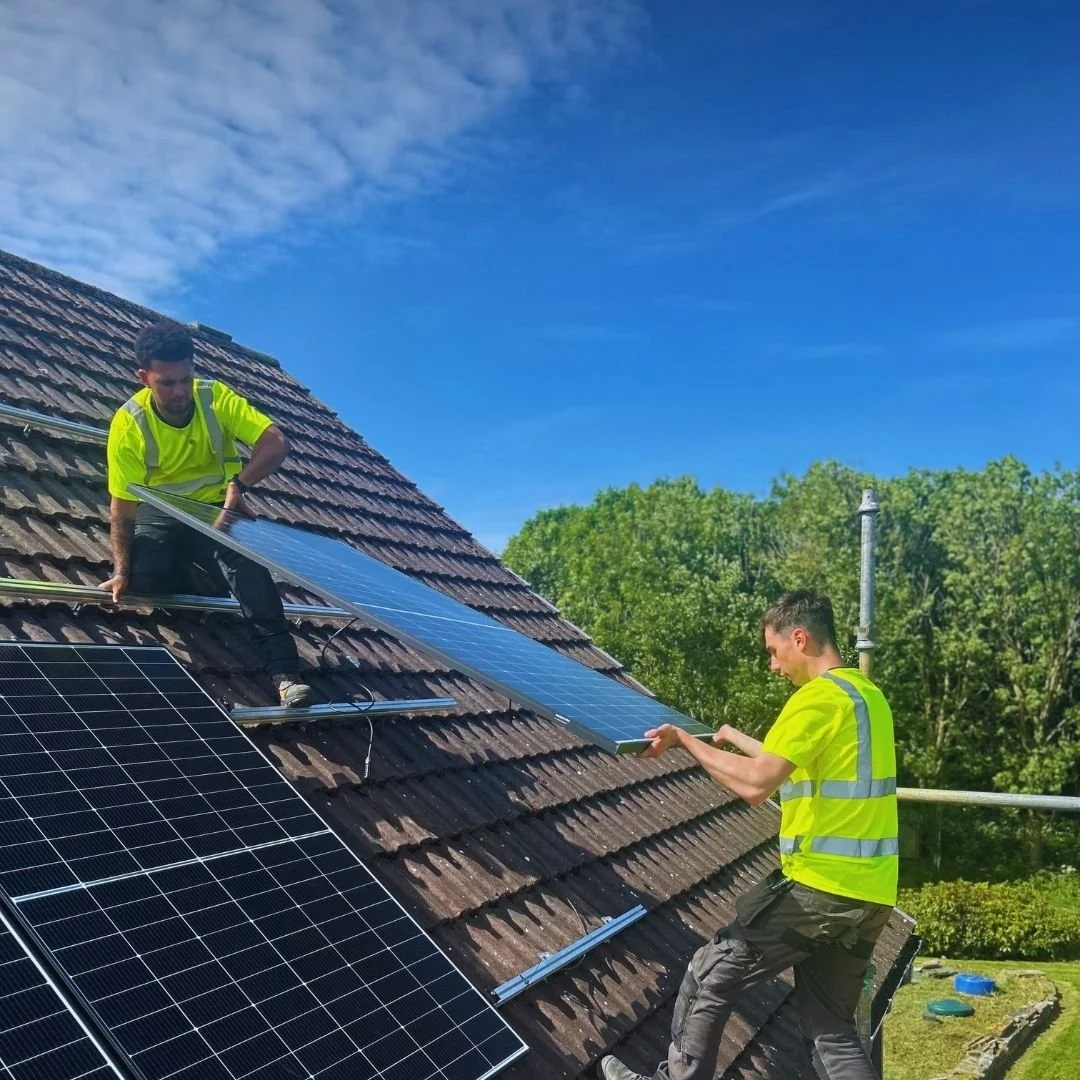 Two workers in yellow safety vests install solar panels on a residential roof with brown shingles, green trees, and a clear blue sky in the background.