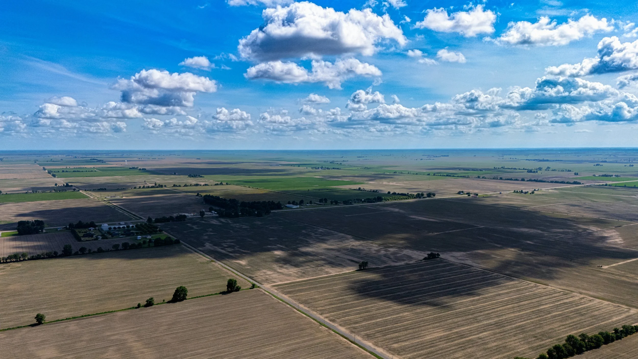 Aerial view of farmland with patchwork fields, scattered trees, and a partly cloudy sky.