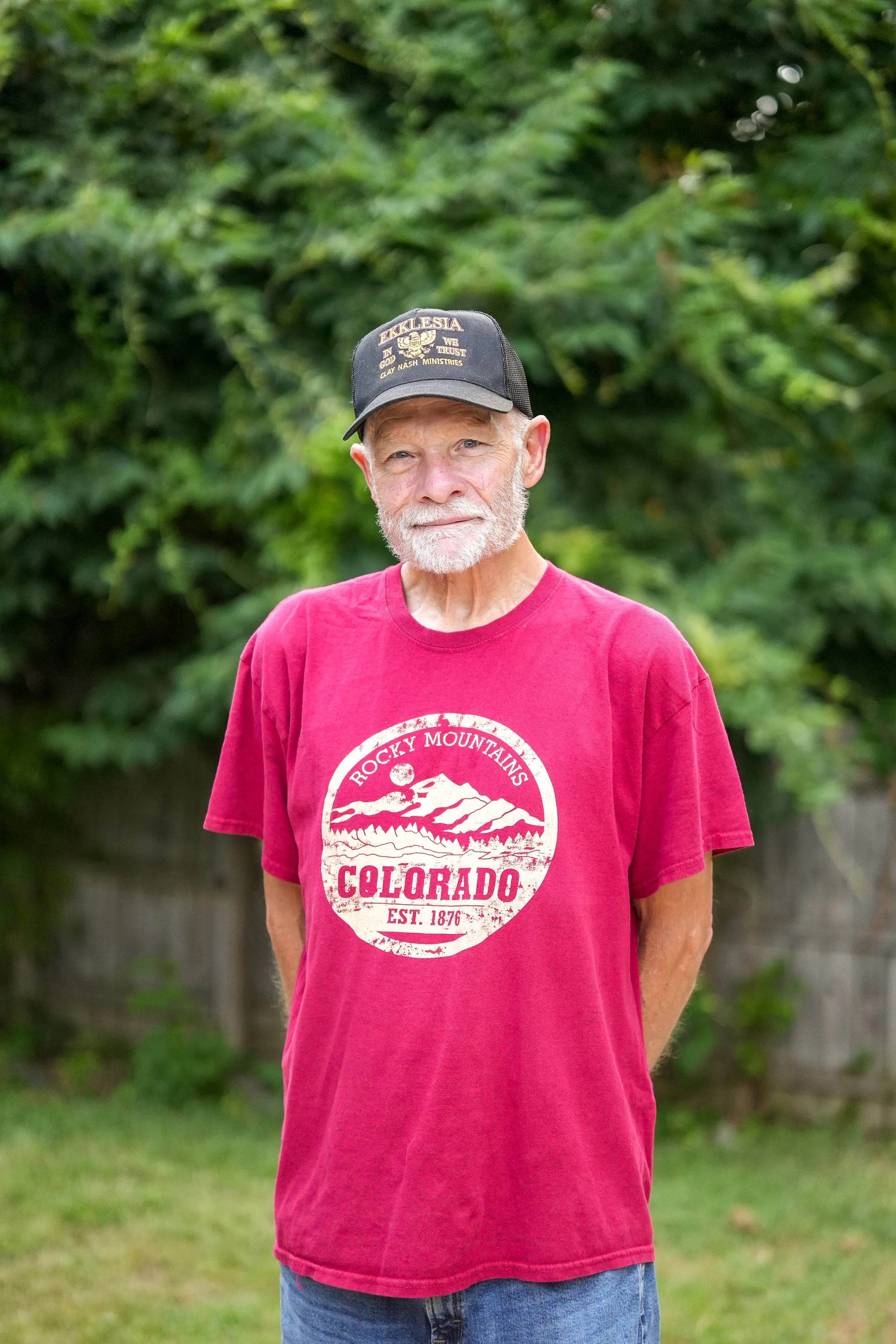 An elderly man with a white beard and mustache, wearing a black cap with yellow text and a red t-shirt with white graphics and text that says 'Rocky Mountains Colorado Est. 1876'. He is standing outdoors with green trees and grass in the background.
