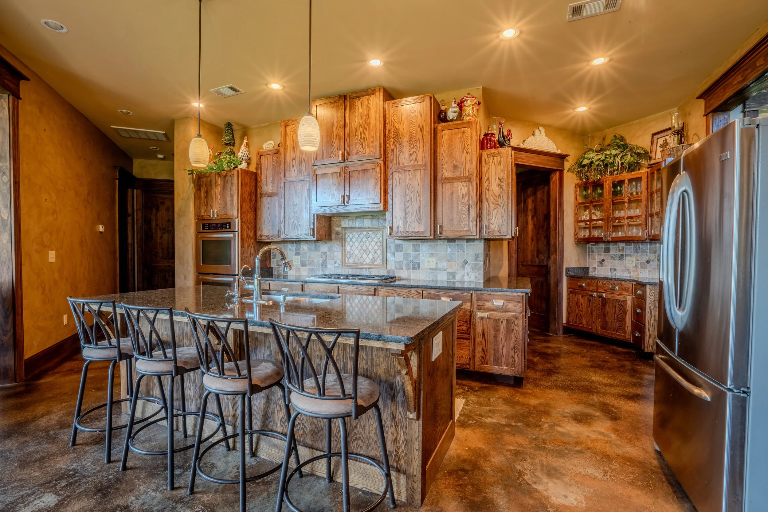 Wooden kitchen with granite island, stainless steel refrigerator, and various cabinets, overhead lighting, and decor on top of cabinets.