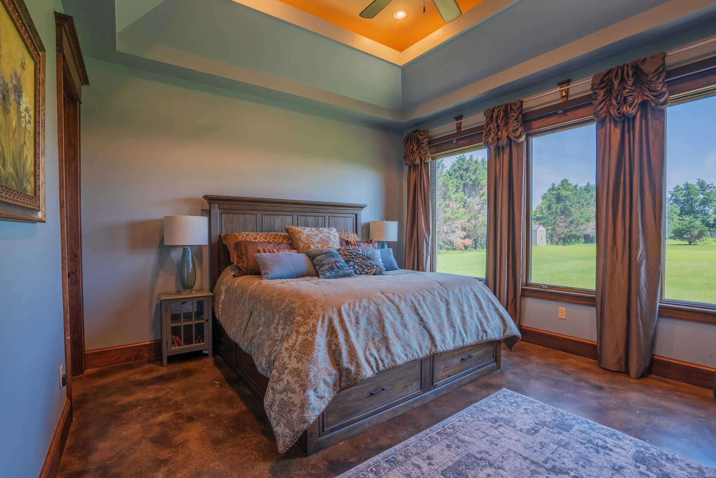 Bedroom with a large bed, multiple pillows, and a wooden headboard. Two bedside lamps on either side of the bed. Large windows with brown curtains overlooking a green lawn and trees outside.
