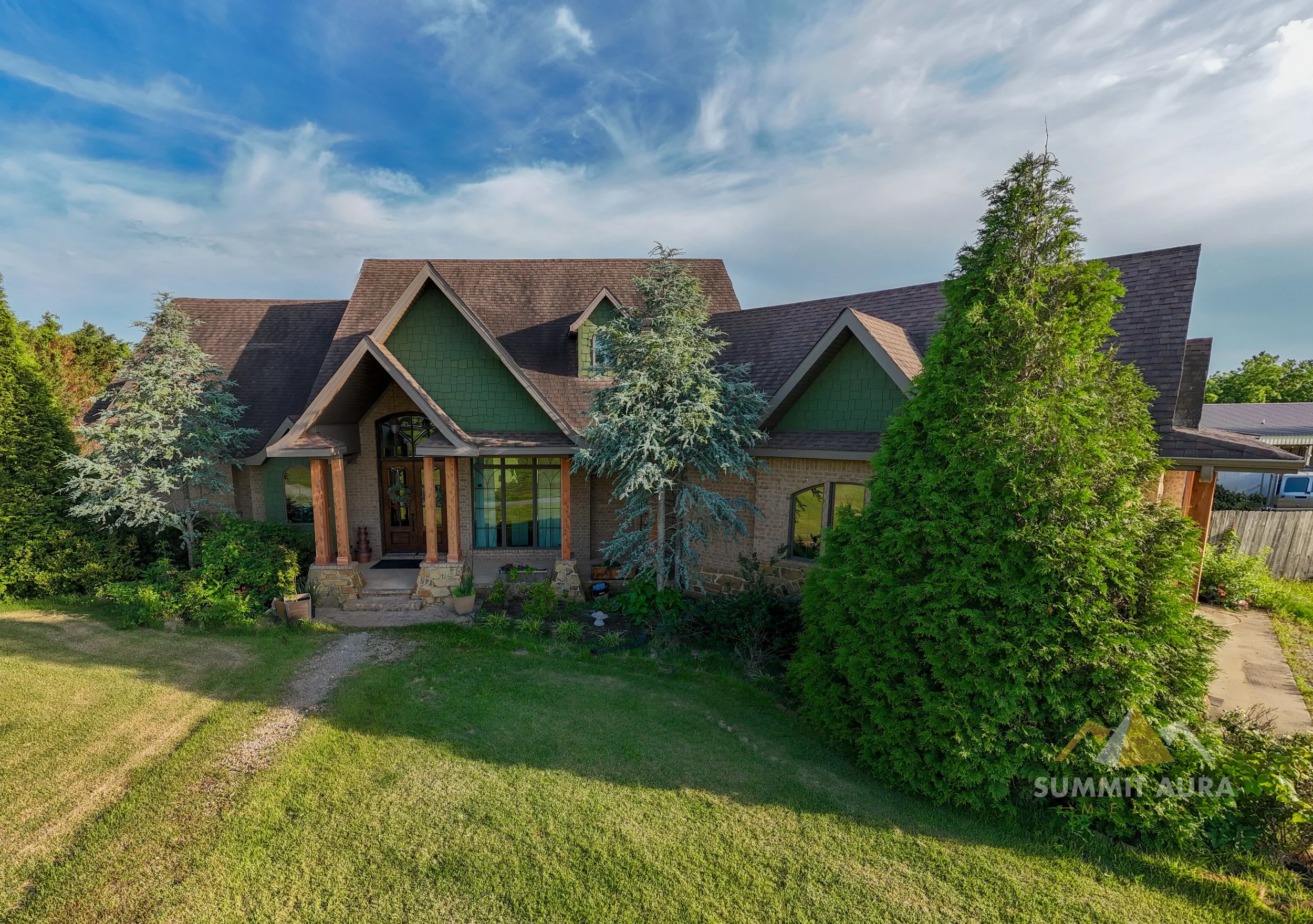A large residential house with multiple peaked roofs, brick and green siding exterior, surrounded by mature trees and a well-maintained lawn, under a partly cloudy sky.