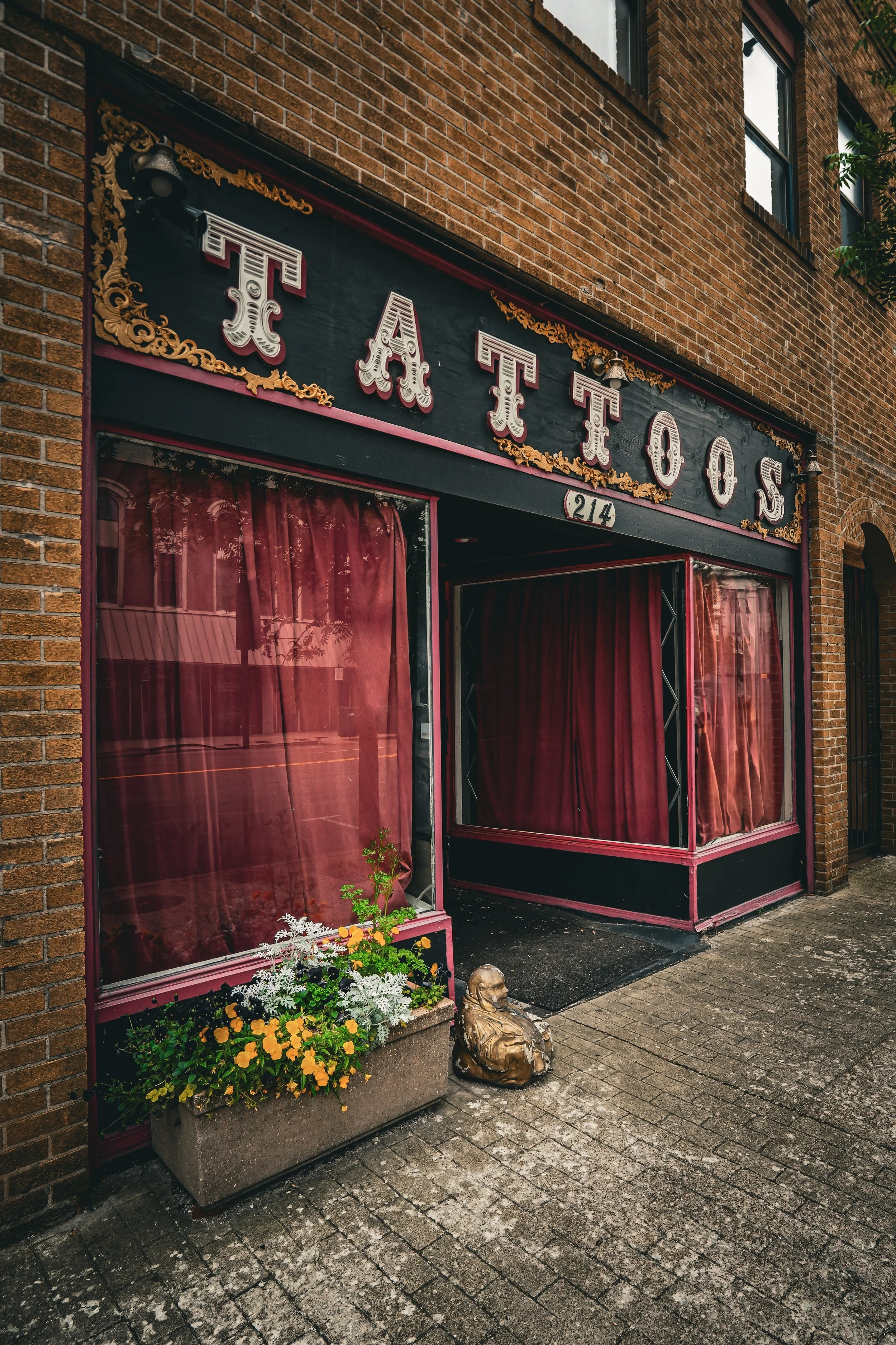 Exterior of a historic theater with a black and red facade, pink curtains, a flower box with yellow and white flowers, and a small Buddha statue outside.