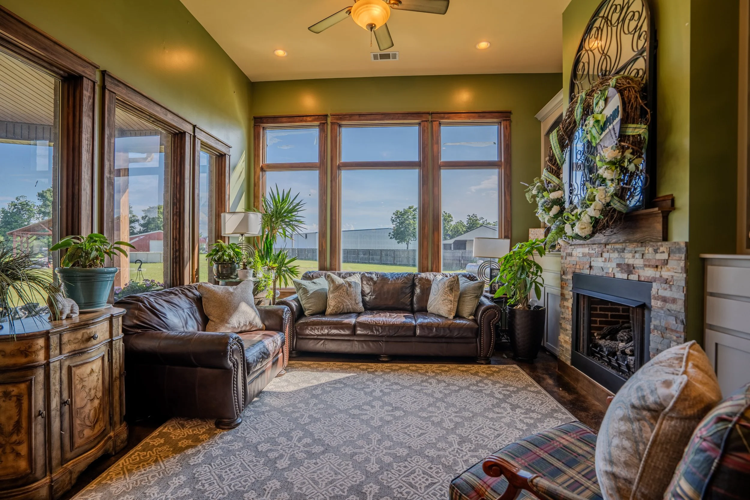 Living room with large windows, leather sofas, potted plants, fireplace with floral wreath, and a patterned area rug.