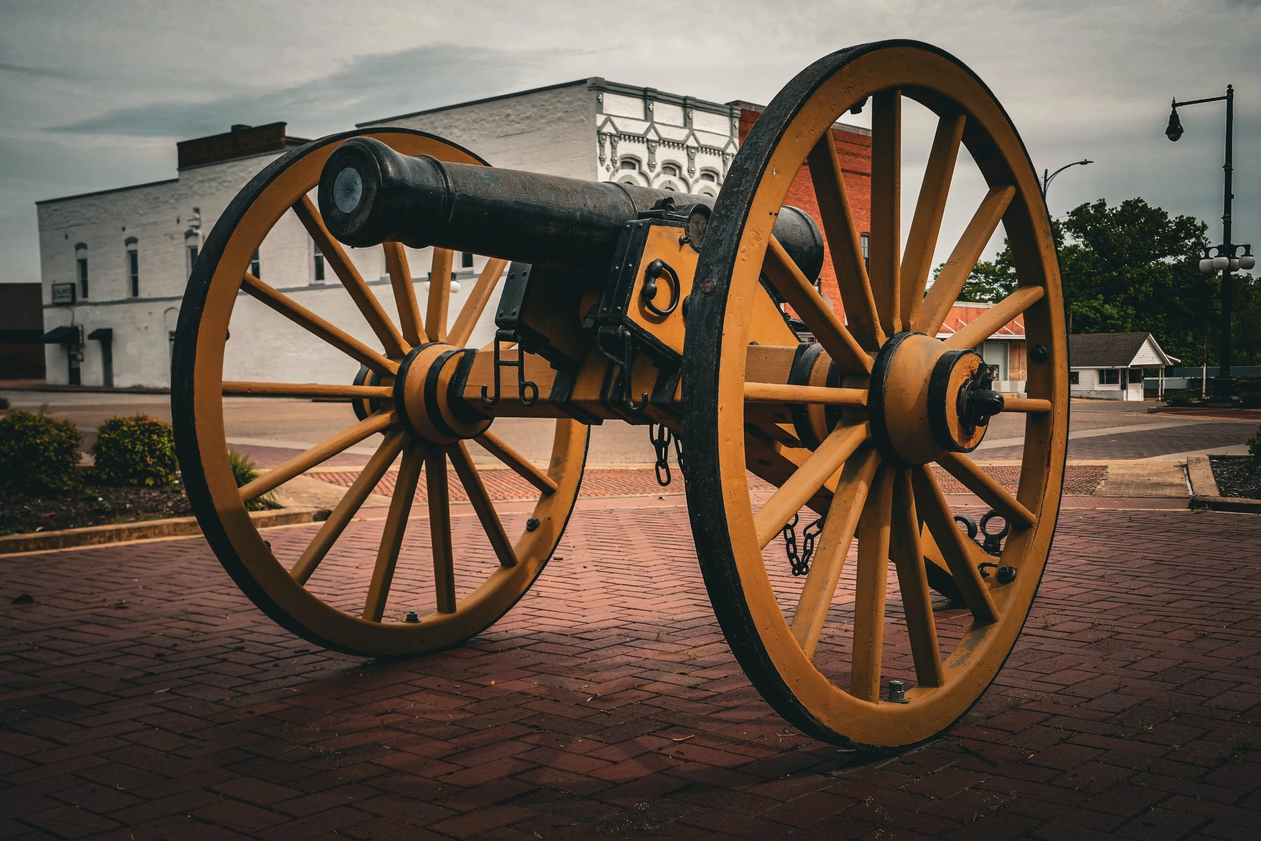 An antique cannon mounted on wooden wheels displayed in a town square with brick pavement, buildings, trees, and street lamps in the background.