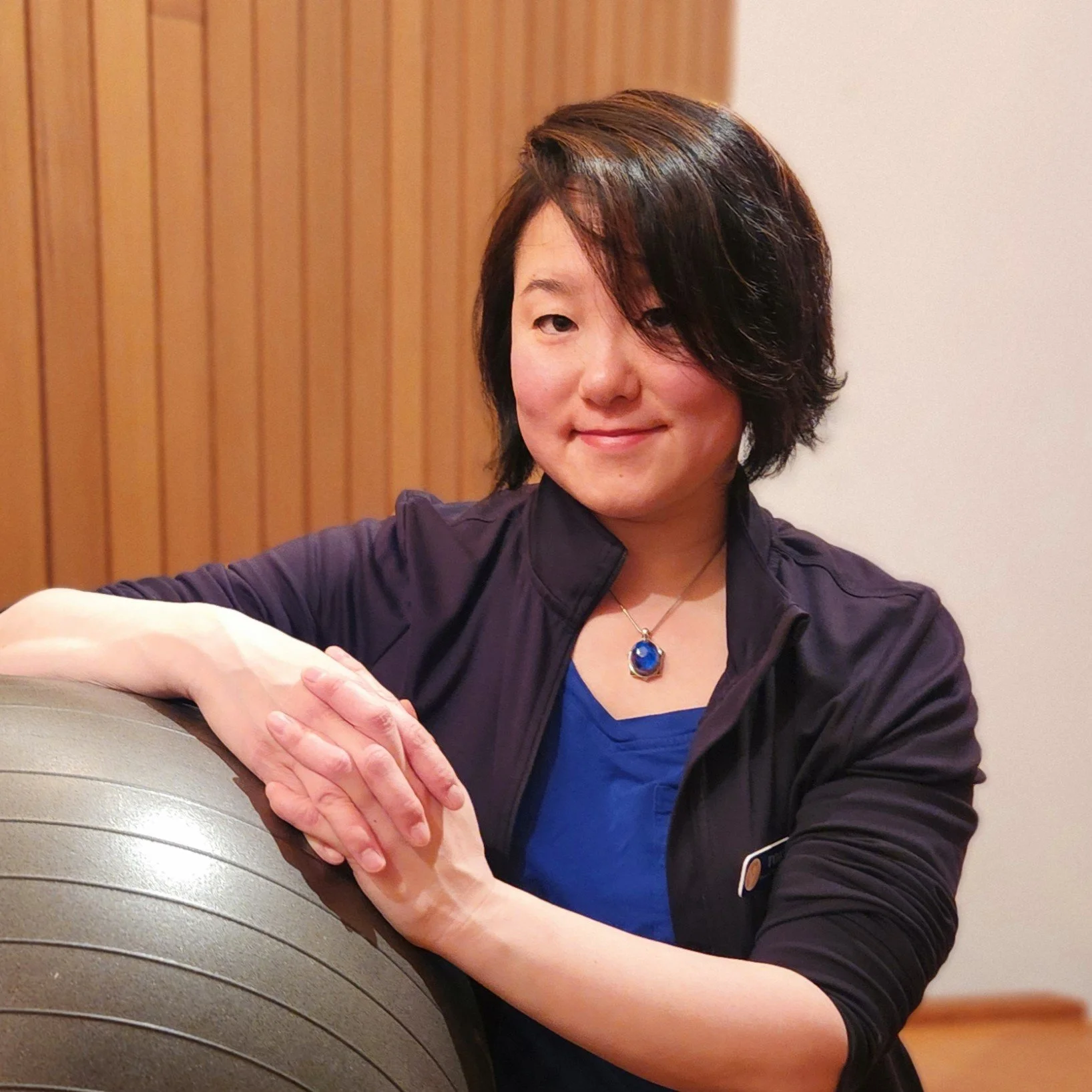 A woman with short dark hair smiling, leaning on an exercise ball, wearing a black jacket, blue shirt, and a blue gemstone necklace.