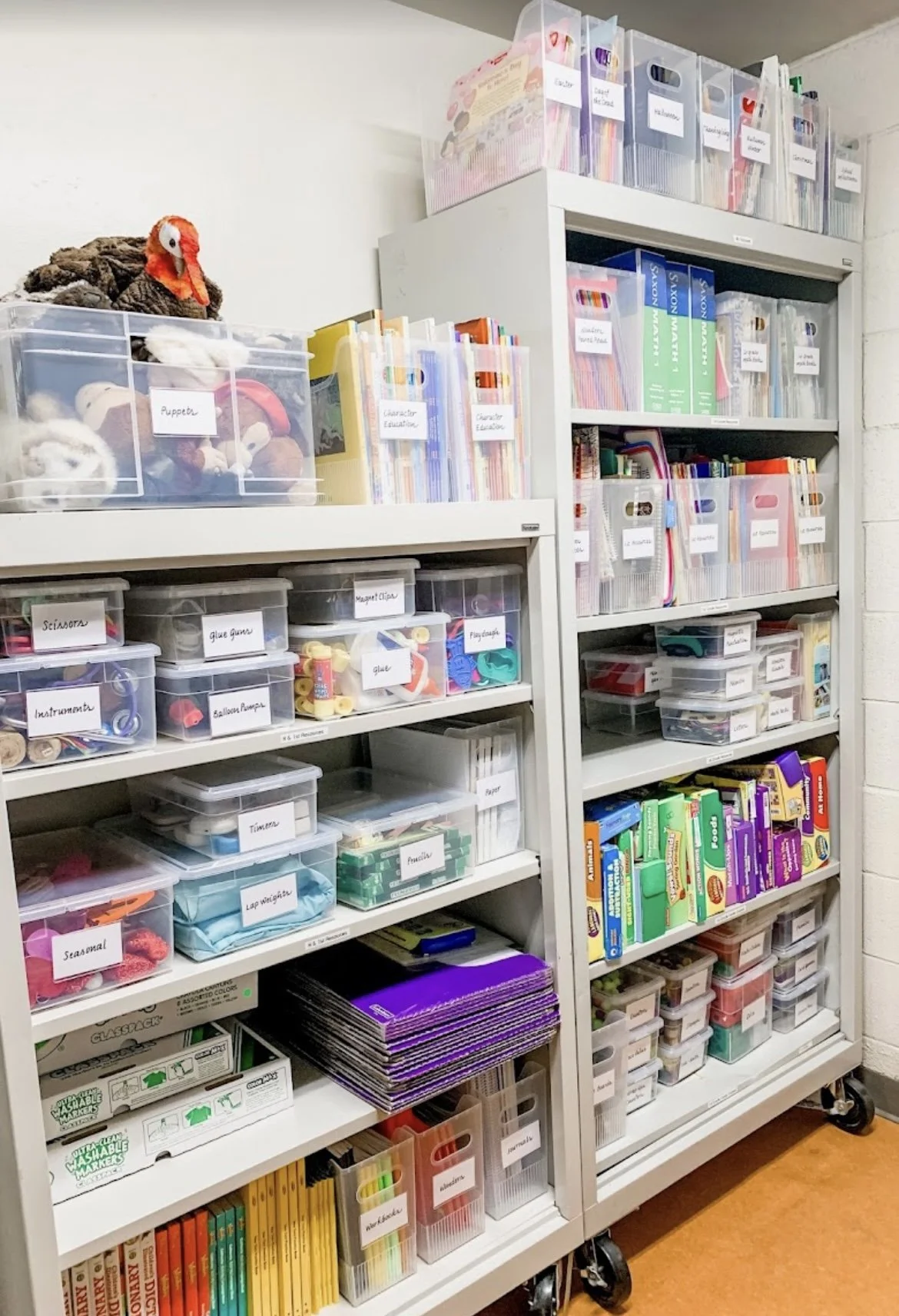 Shelves filled with organized craft supplies including markers, glue, scissors, foam shapes, paper, and crafting materials. Clear labeled boxes and some plush toys or puppets on top.