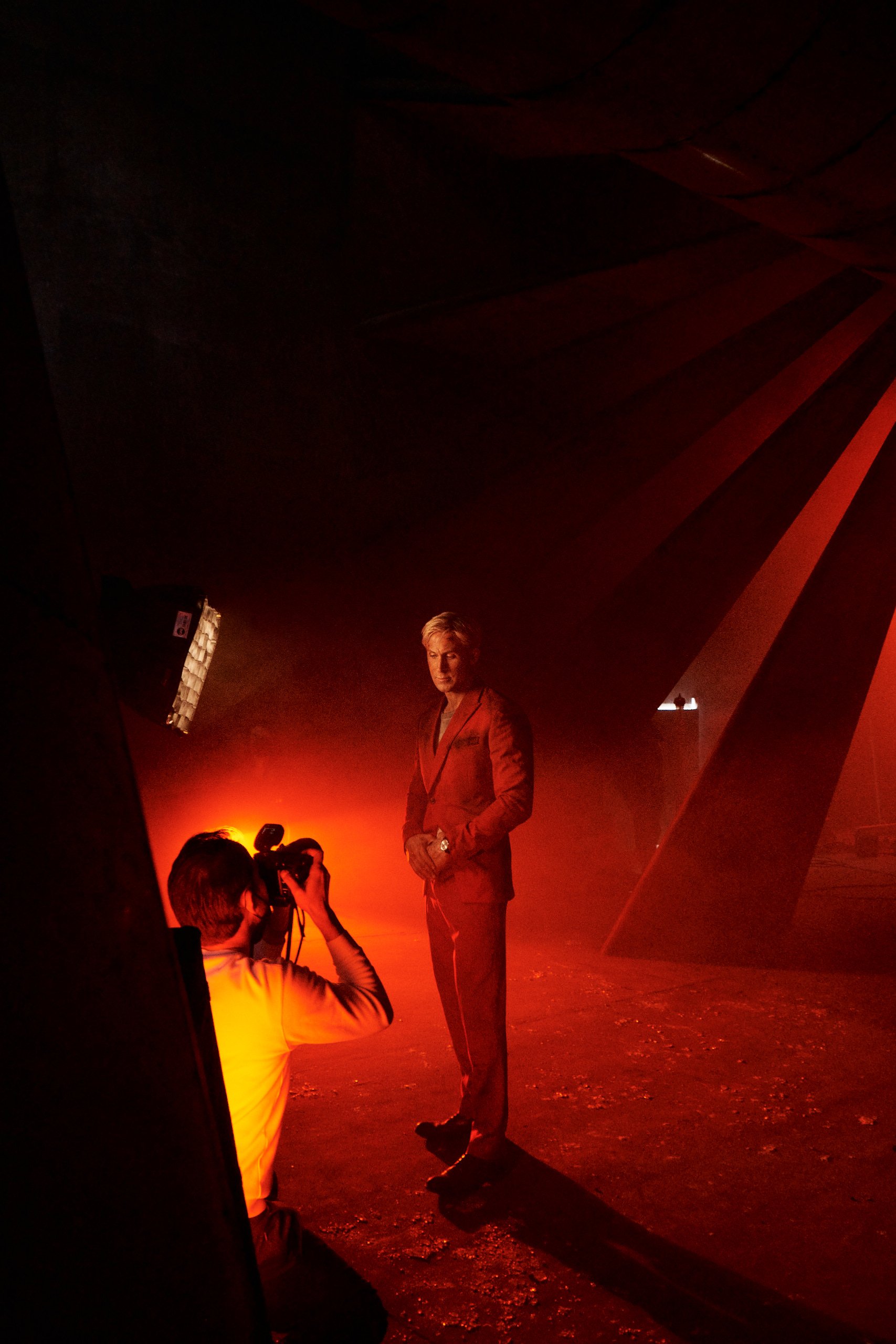 A woman in a red suit standing in front of a photographer who is taking her picture in a dark setting with red lighting and large geometric structures in the background.