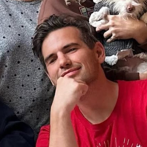A young man with dark hair, resting his chin on his hand and smiling, sitting outdoors with a dog in the background.
