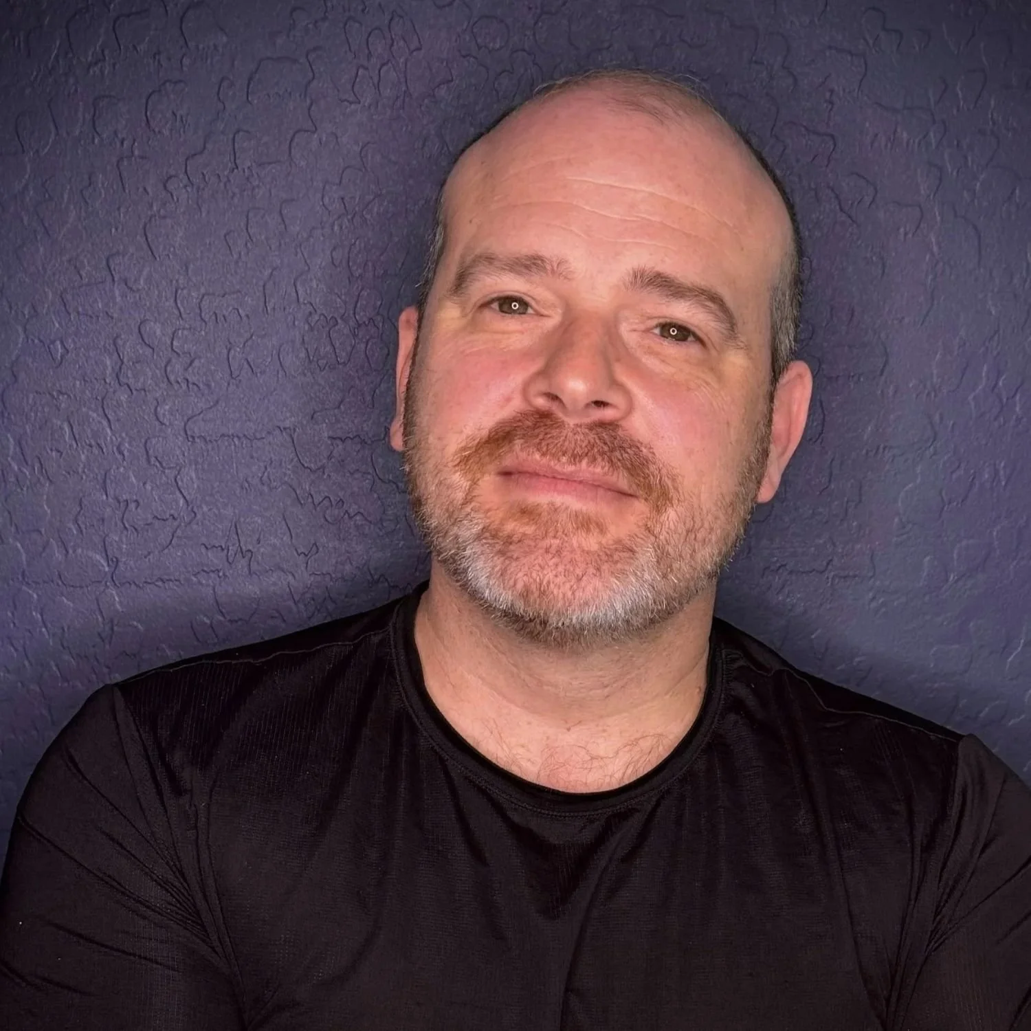 Joel B. Feldman, Founder and Principal Coach of Look Newly, standing with his arms crossed against a dark blue textured wall.