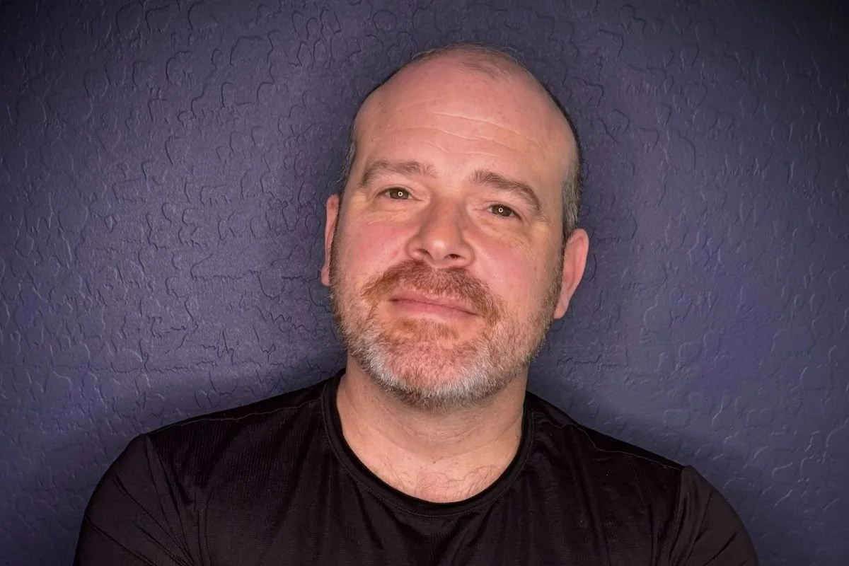 Joel B. Feldman, Founder and Principal Coach of Look Newly, standing with his arms crossed against a dark blue textured wall.
