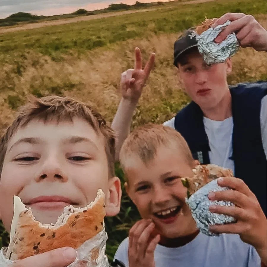 Fem unge drenge, afslappet og leger på en sofa i et kammeratrum. To ligger og chiller, mens en sidder og peger mod kameraet, en ligger med benene oppe, og en ligger og sover. Bag sofaen er en væg med et farvet skilt med et træ og blomster, og teksten 'Må ikke Betrædes!'