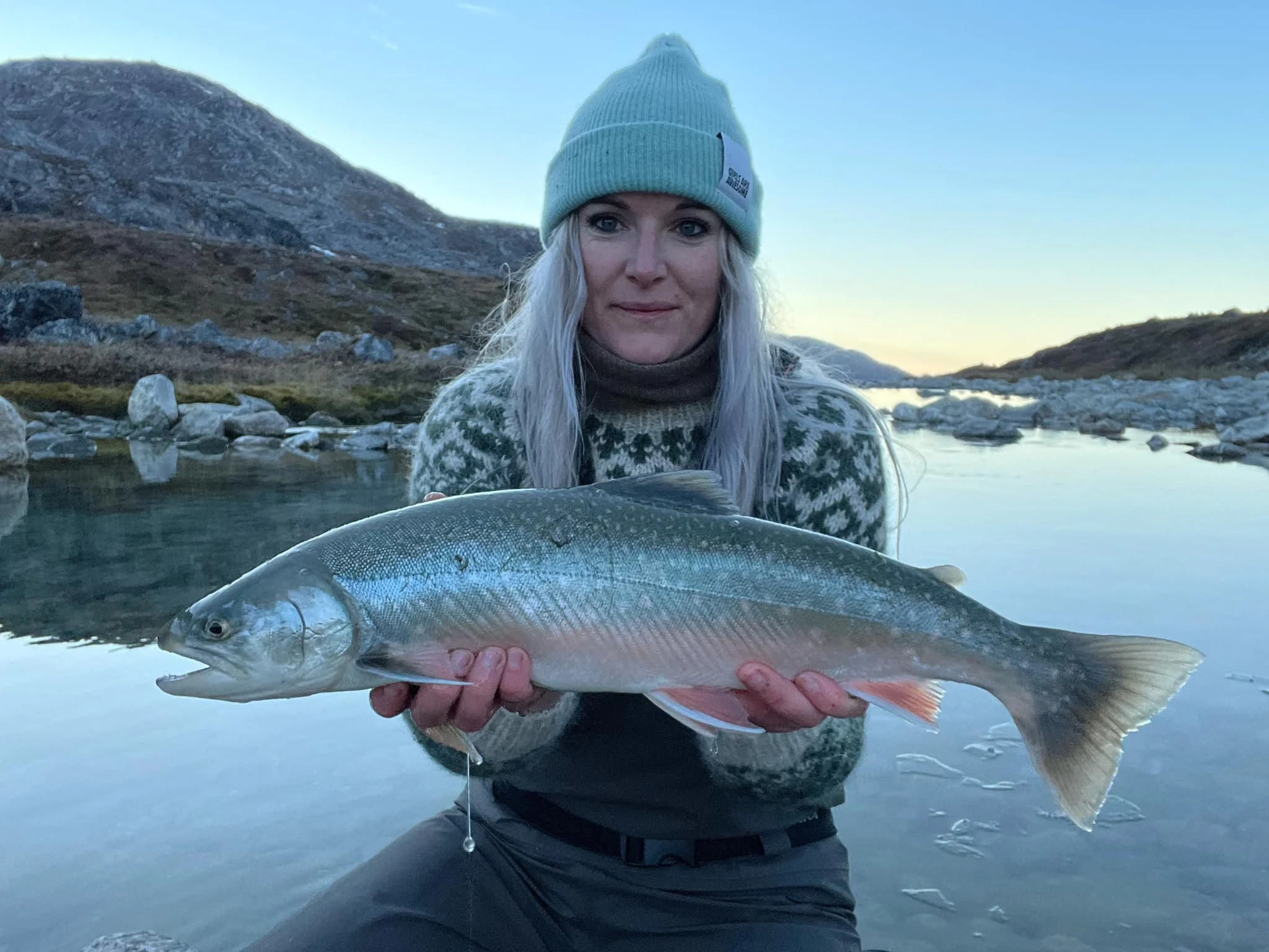 Anne Kjær Lindegaard Eholder en stor fisk foran en naturskøn flod med bjerge i baggrunden. Det er vinter med sne på jorden. Hun fortæller om hvorfor hun er frivillig i en lystfiskerforening.