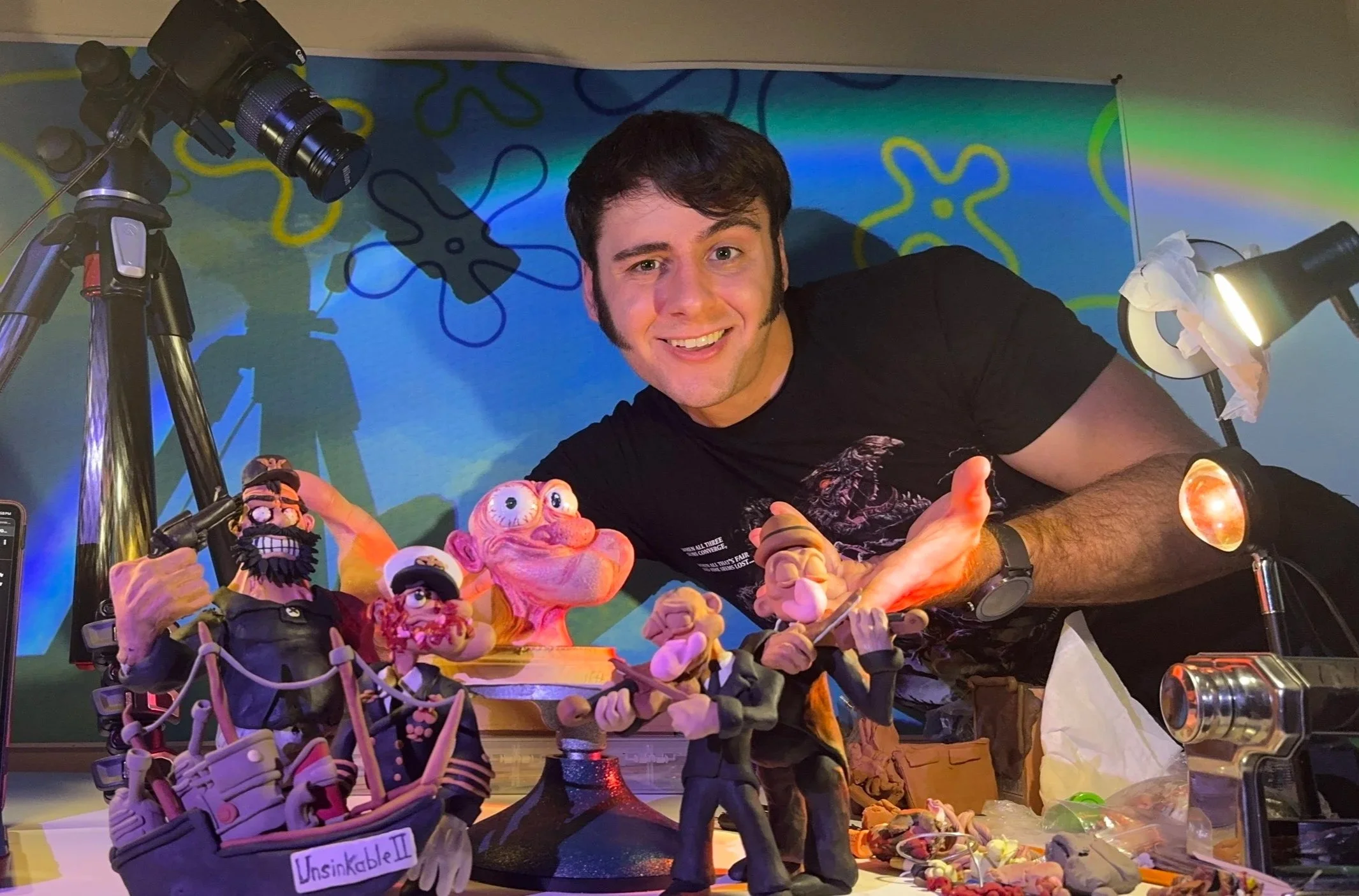 A smiling man with dark hair and a beard crouches behind a table filled with various colorful puppets and figurines, with a backdrop of a blue and green abstract mural. There is a camera on a tripod and lighting equipment nearby.
