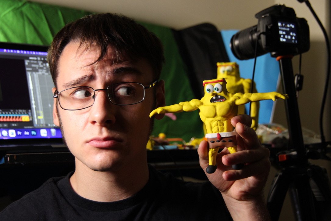 Young man with glasses holding a yellow muscular SpongeBob SquarePants puppet with a surprised expression, in a room with photography equipment and a computer in the background.