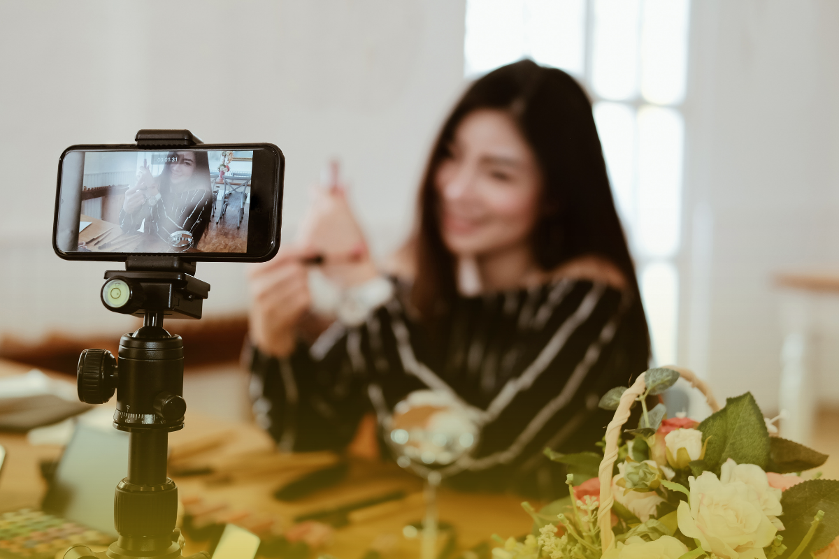 A woman films a video. Her phone is on a tripod, and she's holding up a product.