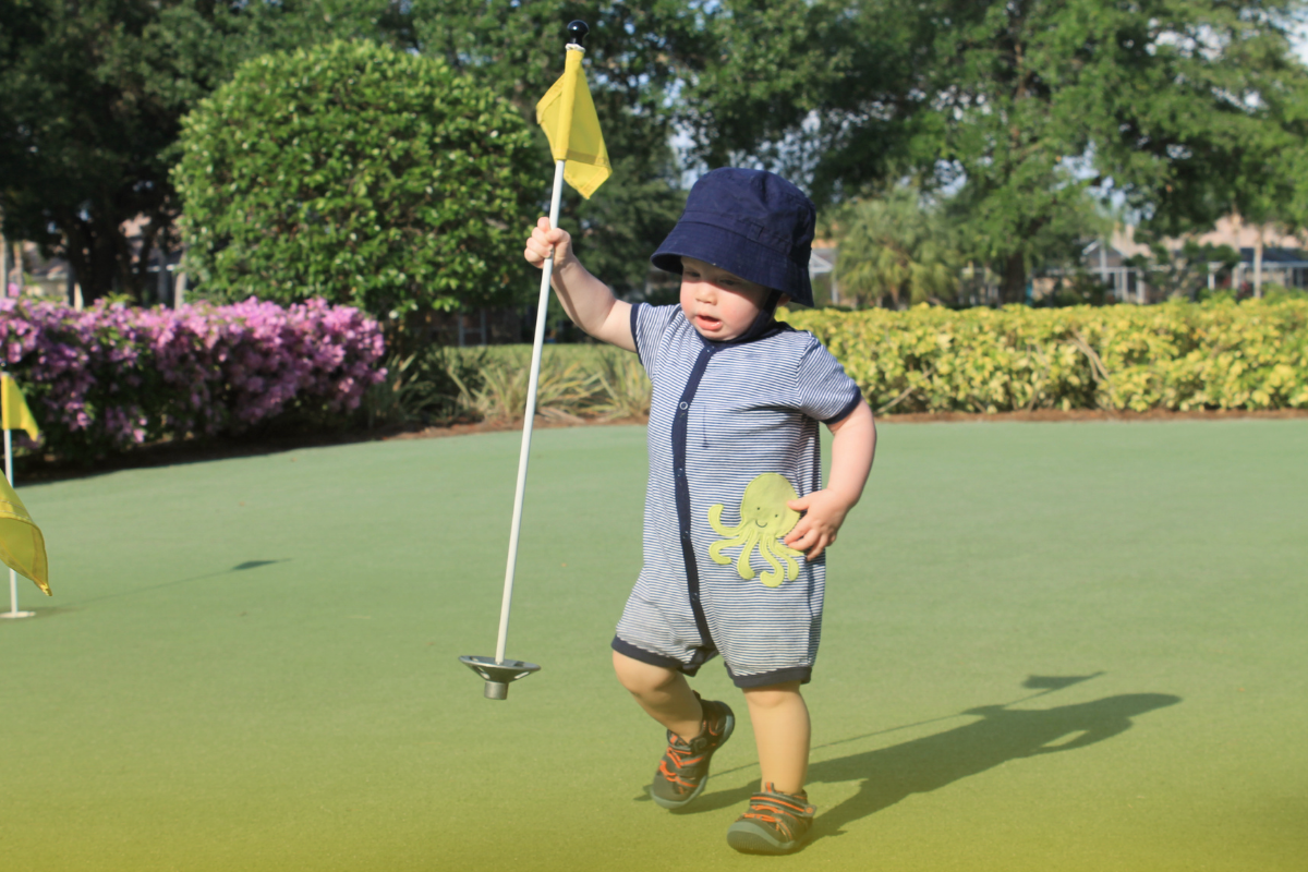 A baby holding a golf flag while standing on a green.