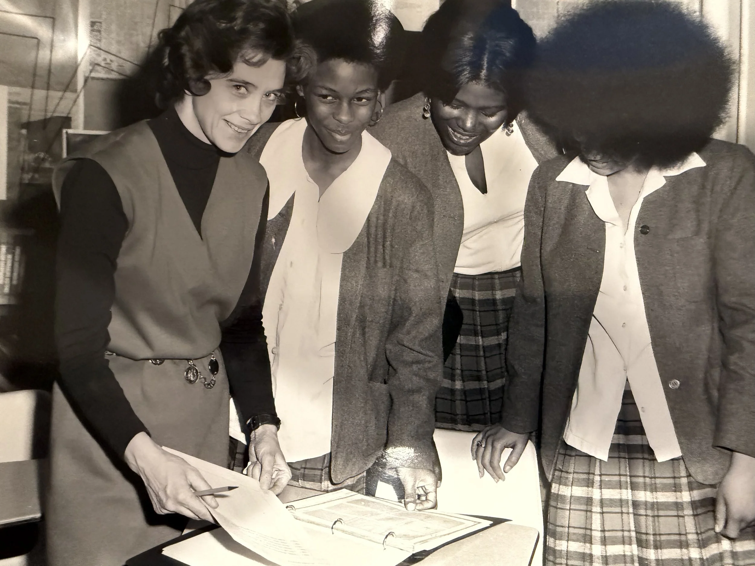 A black and white photo of four young women gathered around a binder