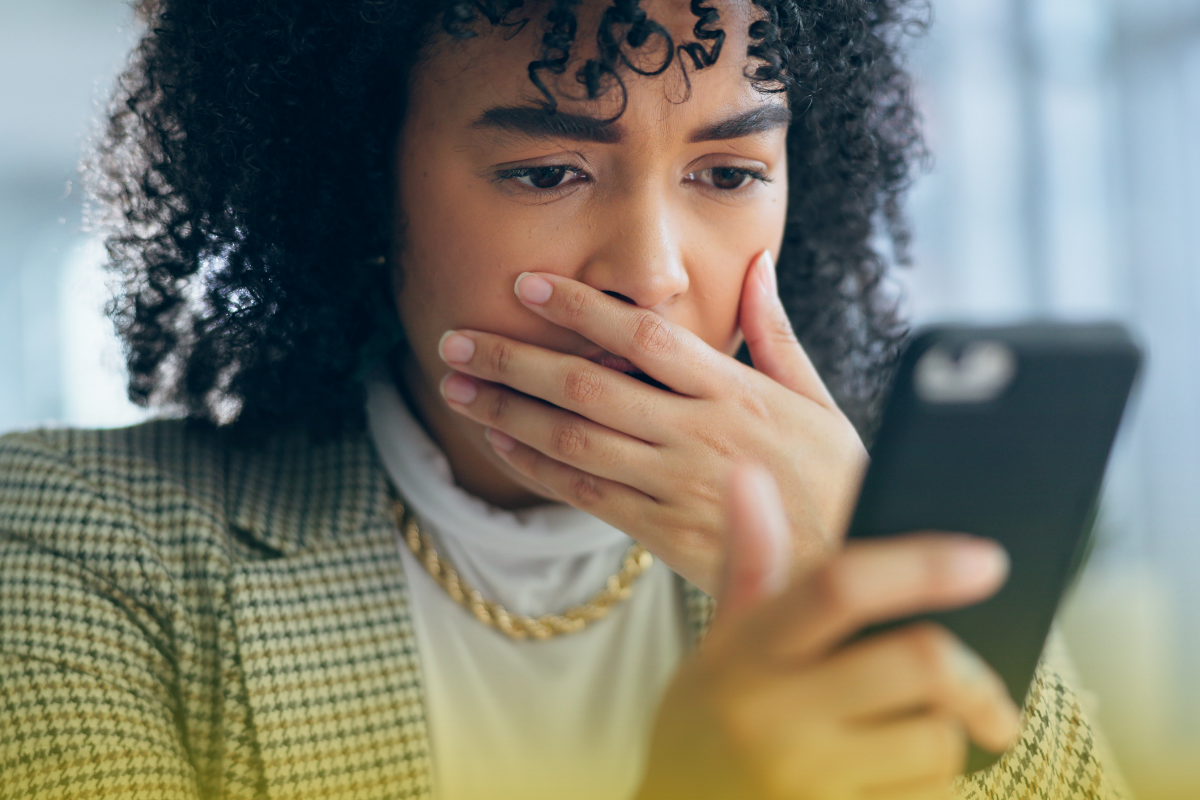 A woman stares at her phone with a horrified expression. She covers her mouth with her hand.