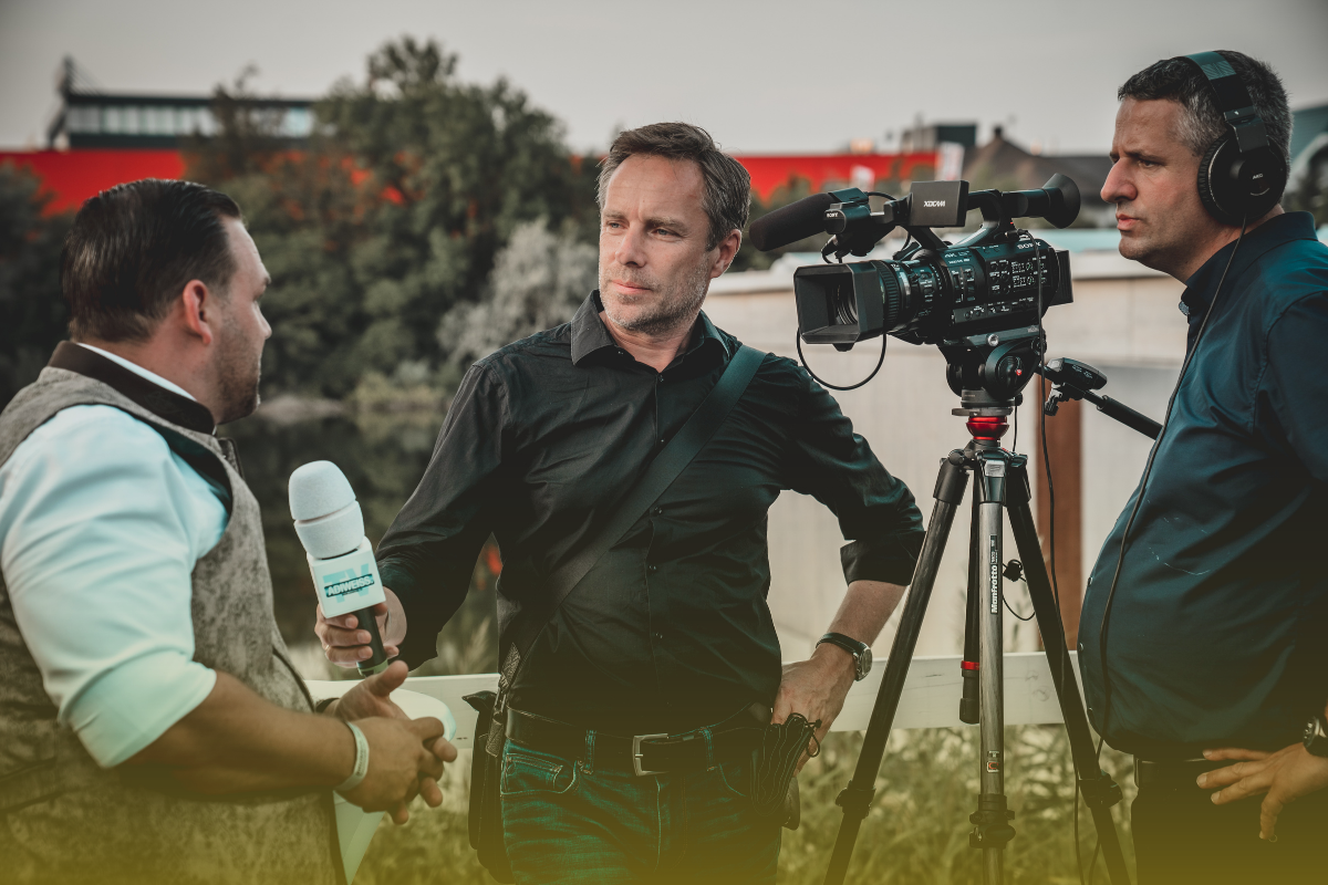A camera man stands filming outside. A male reporter hands out a microphone for an interviewee to speak into.