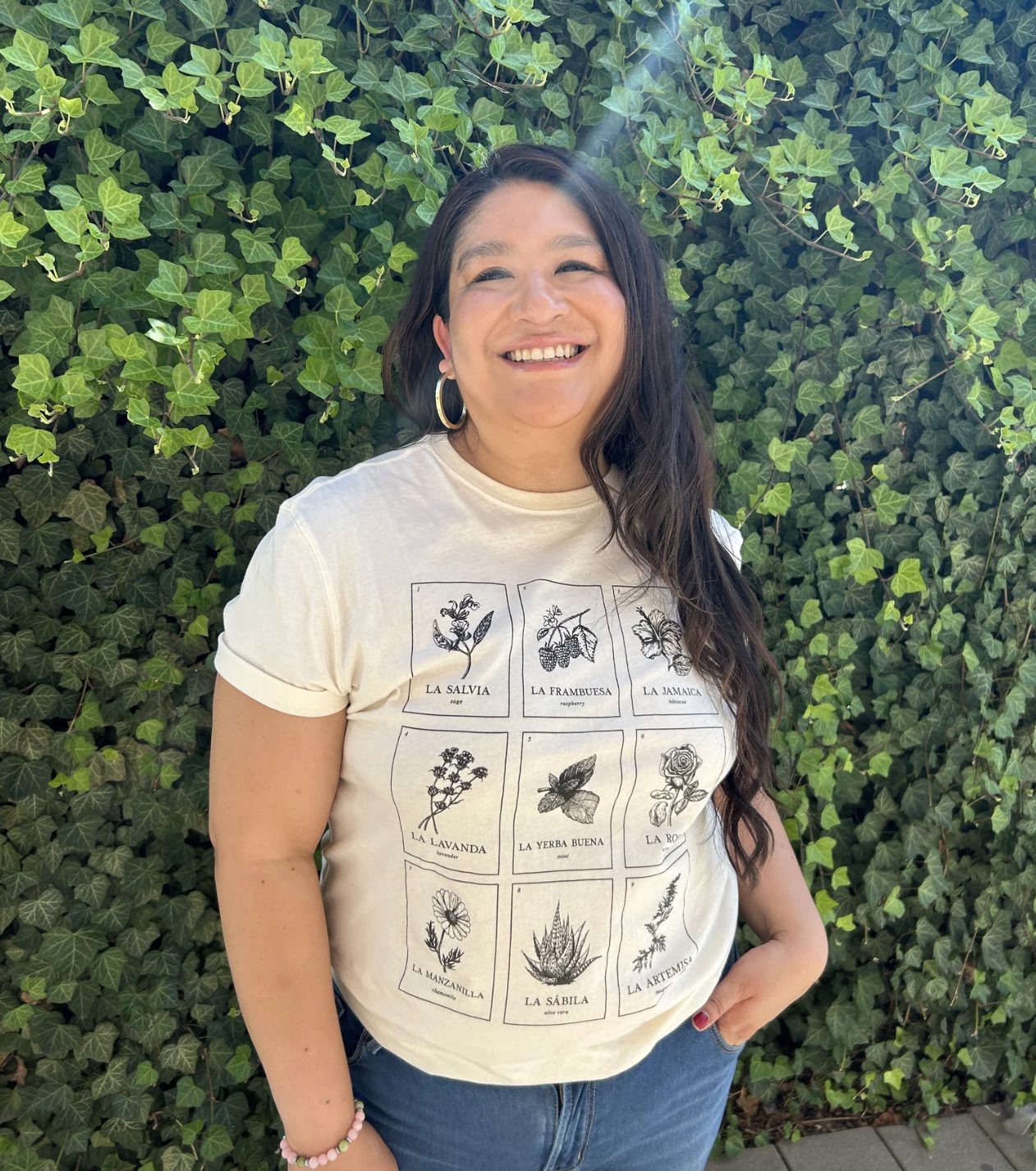 Smiling woman with long dark hair wearing a beige loteria style T-shirt with illustrations and names of various herbs, standing in front of a lush green ivy-covered wall.