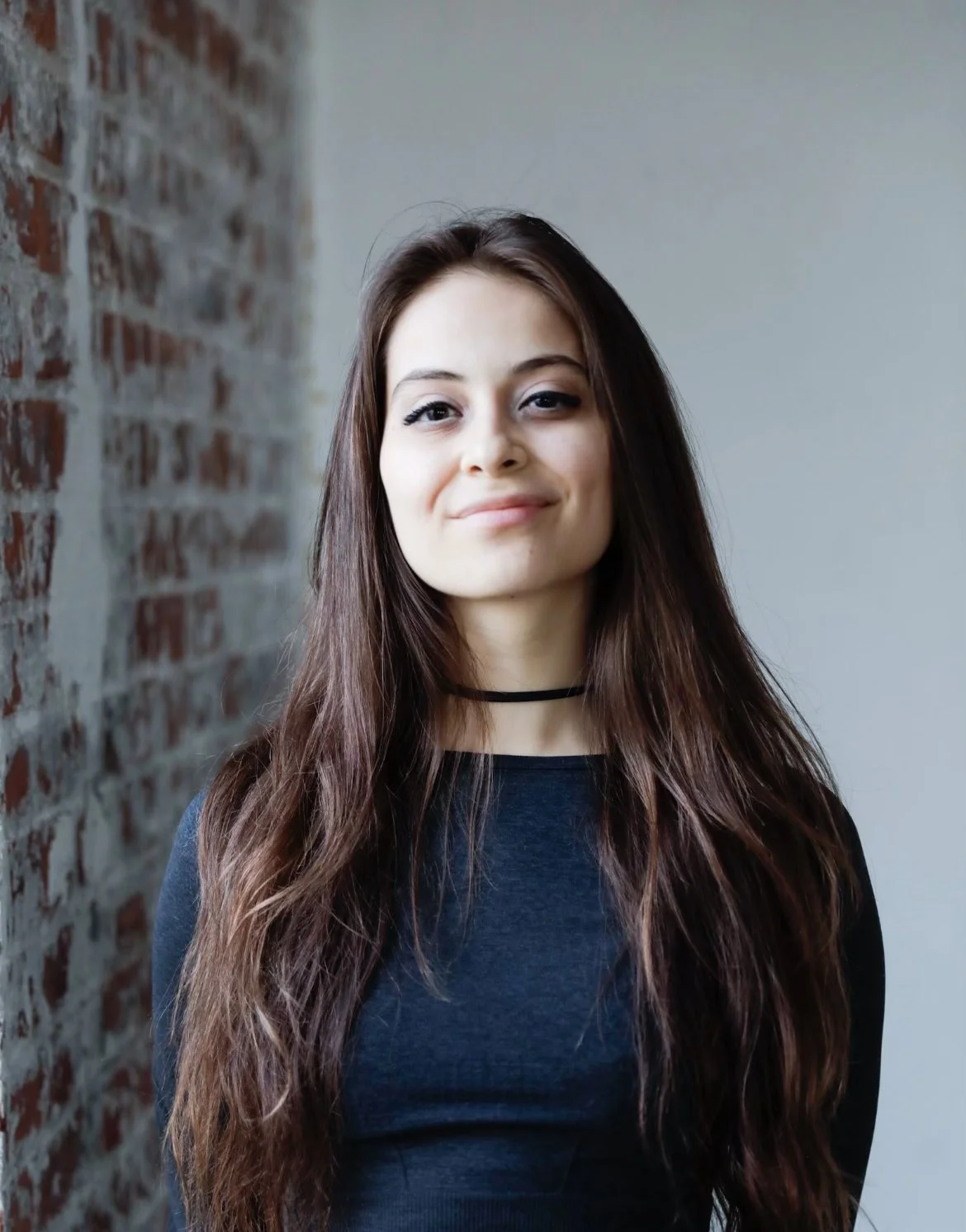 A young woman with long brown hair, wearing a black choker and black top, standing indoors next to a brick wall, smiling slightly.