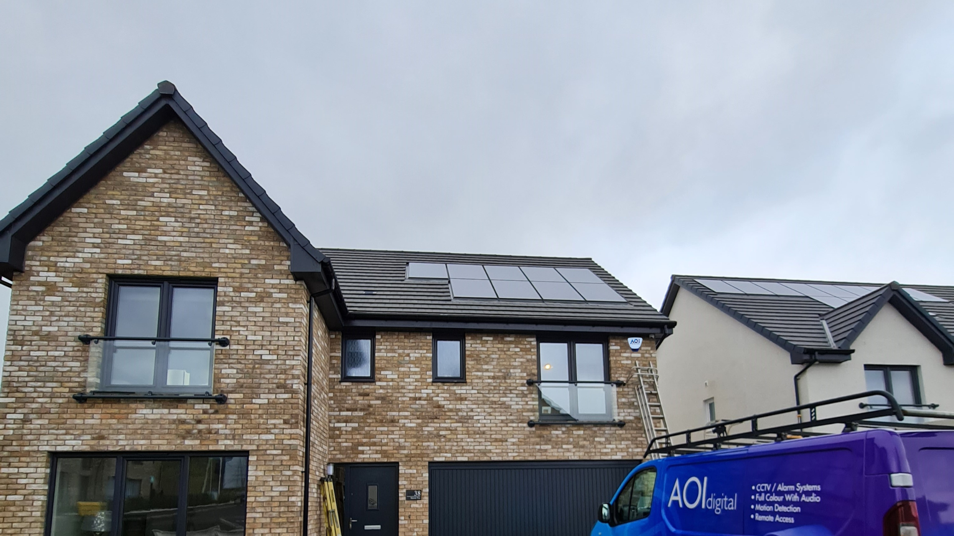 Modern brick house with solar panels on the roof, a glass balcony, and a blue security system service van parked in front.