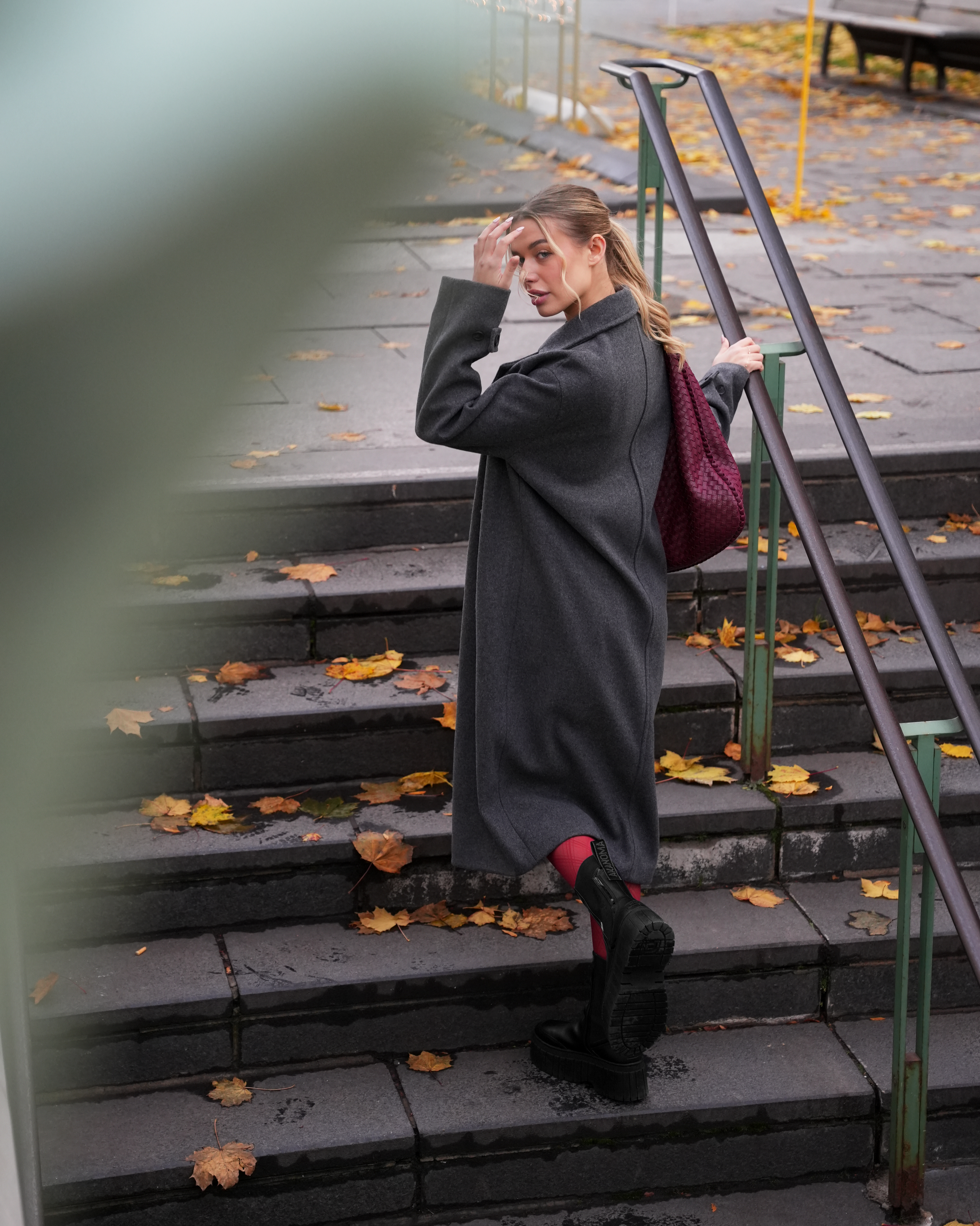 Woman in a long dark coat standing on outdoor steps covered with autumn leaves.
