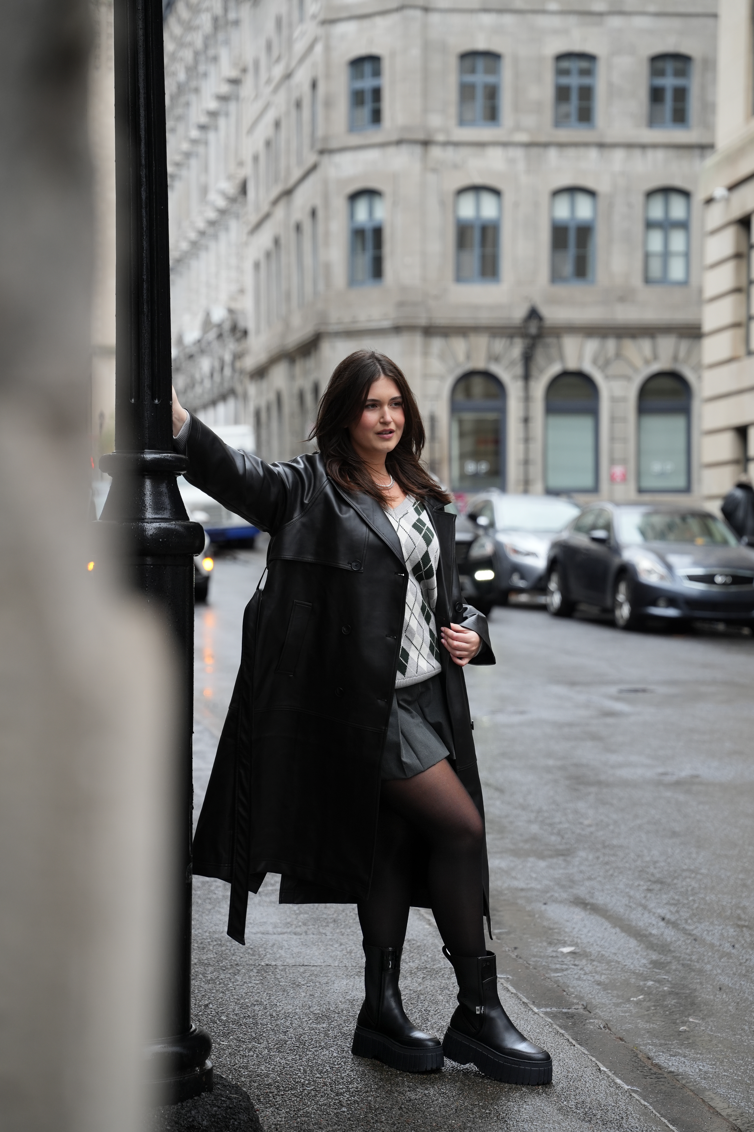 Woman in a dark coat posing beside a building on a wet city street.