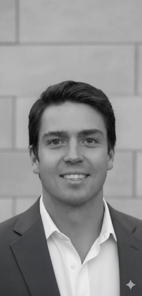 A young man with short, neatly combed hair, wearing a suit and a collared dress shirt, smiling at the camera in front of a plain light-colored wall.