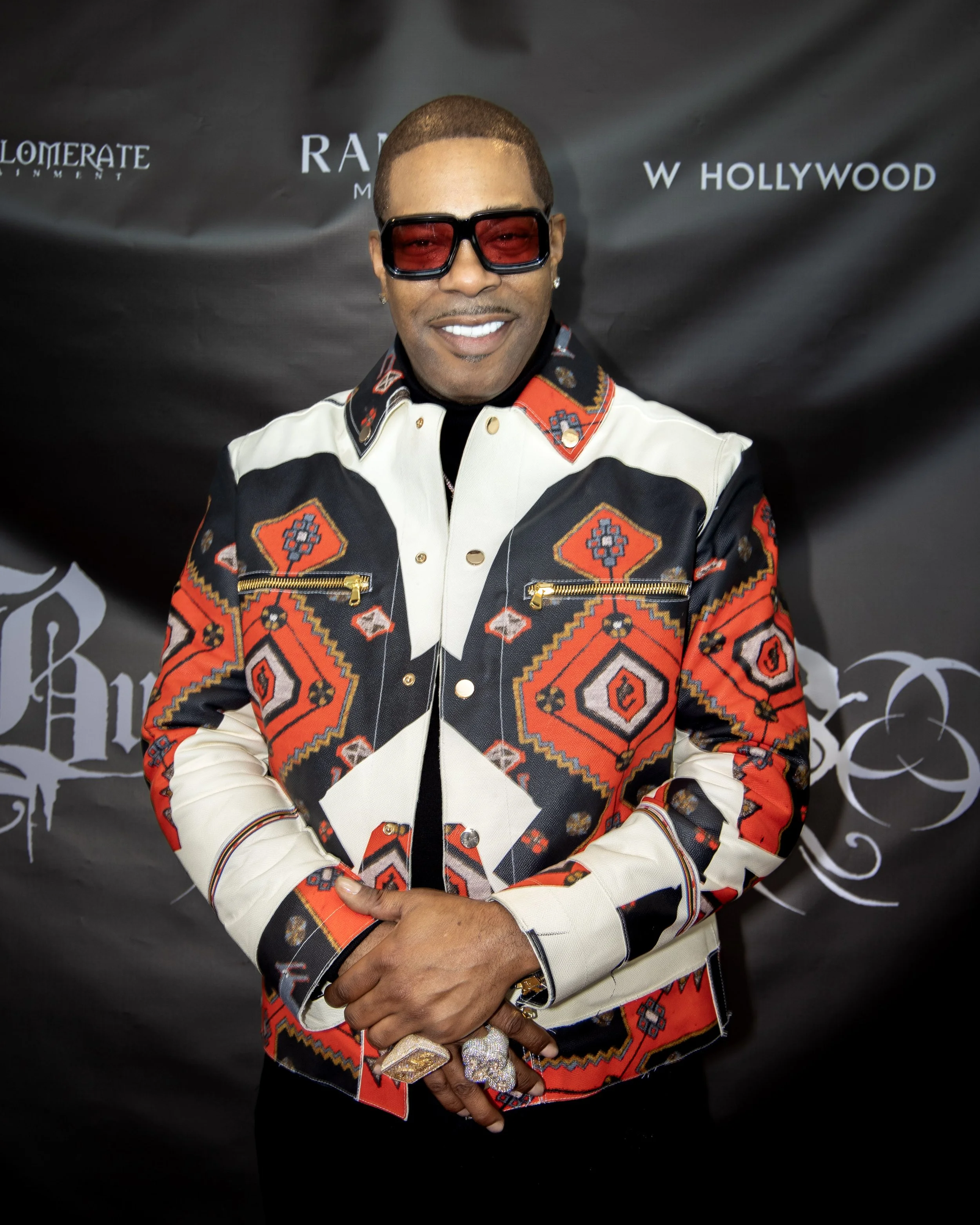 Photo of a man with short hair wearing red-tinted glasses and a colorful patterned jacket, smiling with his hands crossed in front of him, standing against a black background.