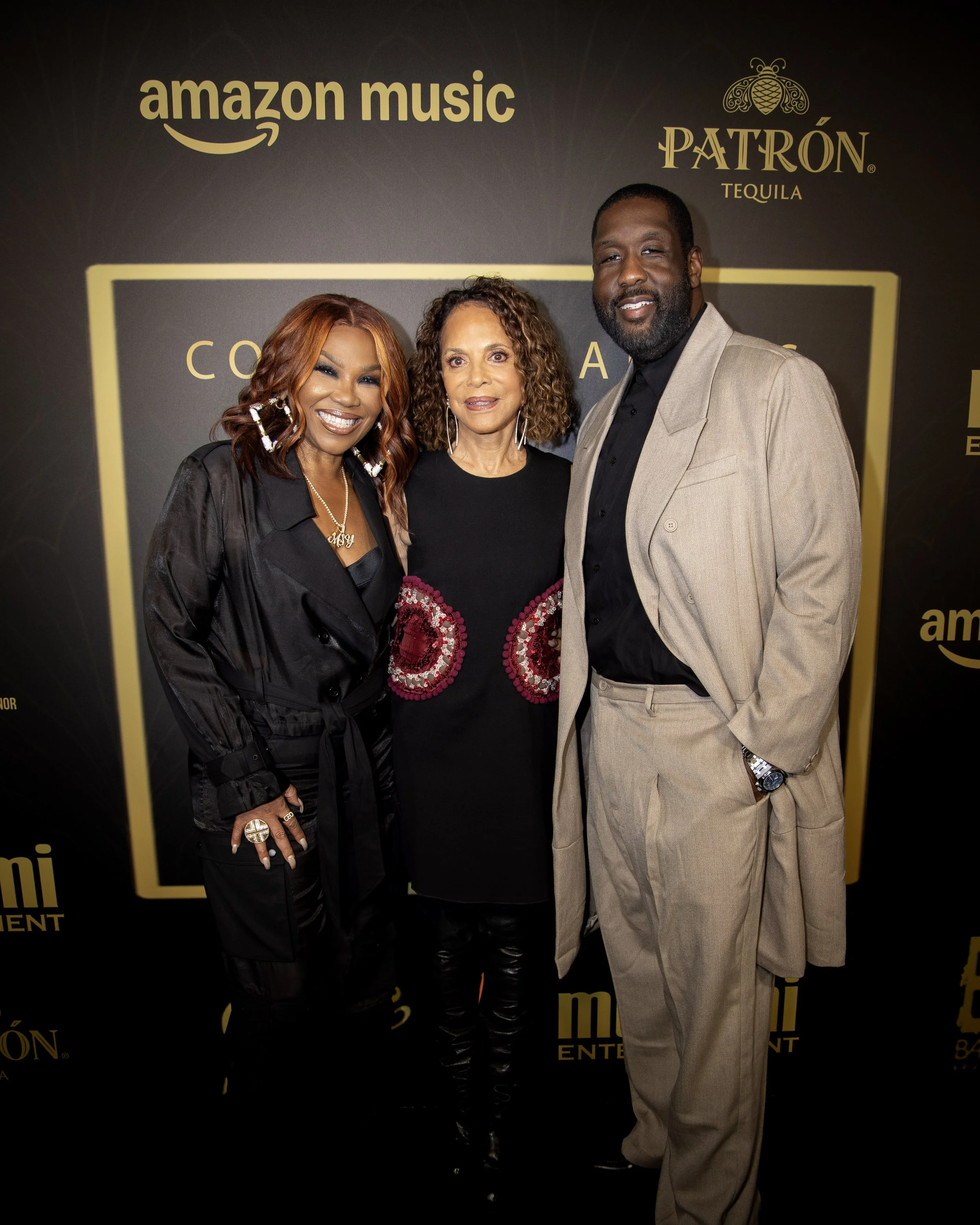 Three people standing together at an event with a black background displaying 'amazon music' and 'Patrón Tequila' logos. Two women and one man, all dressed in formal attire, smiling and posing for the photo.