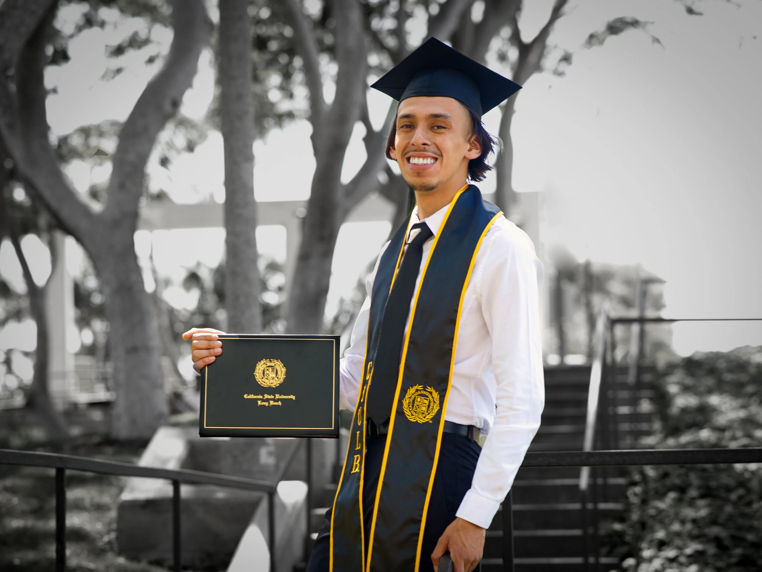 A young man in graduation attire, wearing a gown, tassel cap, and honor cords, holding a diploma folder from California State University, standing outdoors with trees in the background and smiling.