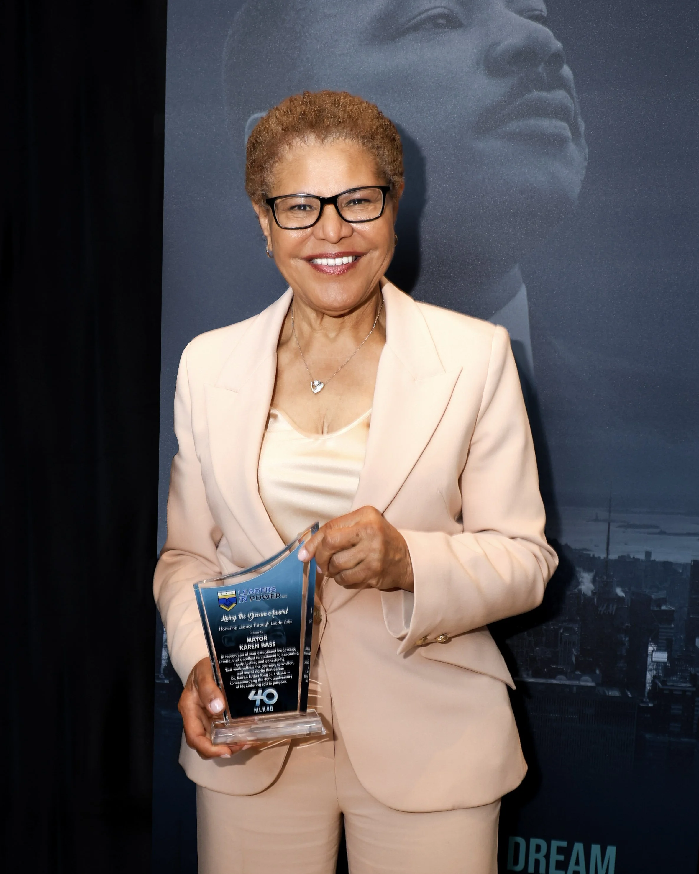 Woman in a beige suit holding an award with a cityscape background. The woman has short curly hair, glasses, and is smiling. The award has the number 40 and the letters MLK40 on it.