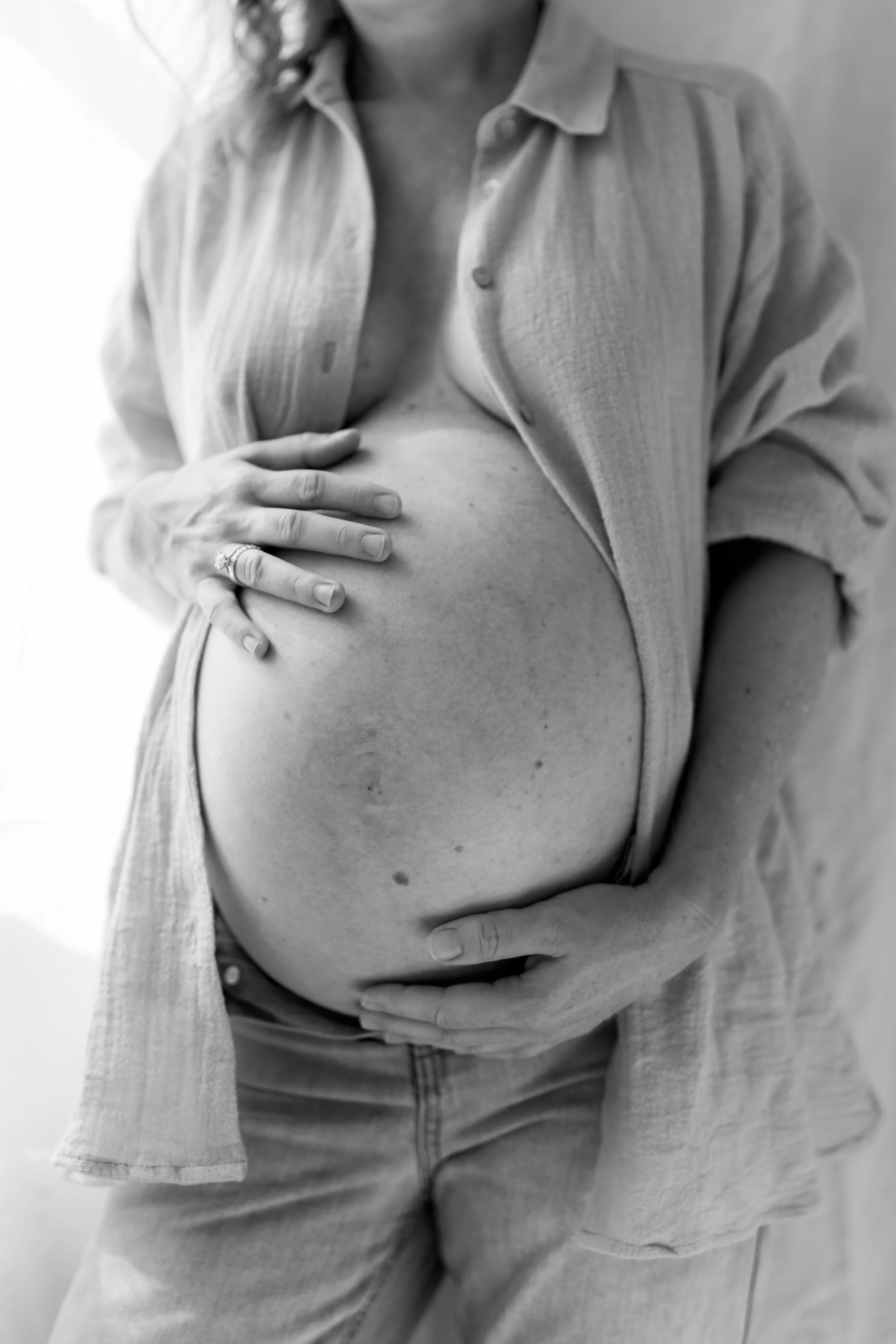 Close-up black and white photo of a pregnant woman showing her belly, with one hand resting on it and the other hand touching her side, wearing a partially unbuttoned shirt and jeans.